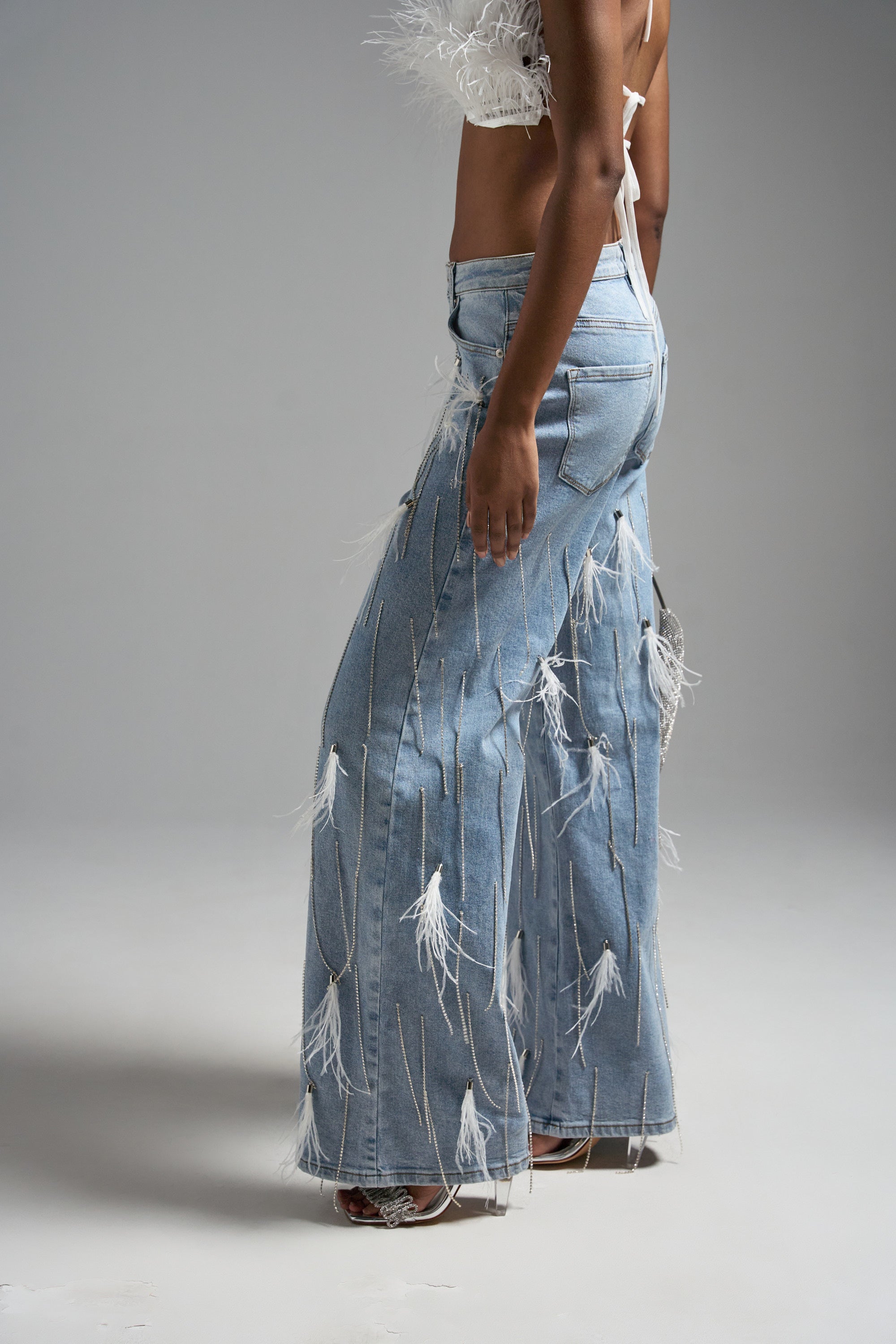 Wearing the SHAKE IT FOR ME GIRL FEATHER EMBELLISHED DENIM—a feathered crop top and high-rise relaxed fit jeans in light blue—a person stands against a plain gray background, with the focus on the jeans and midsection.