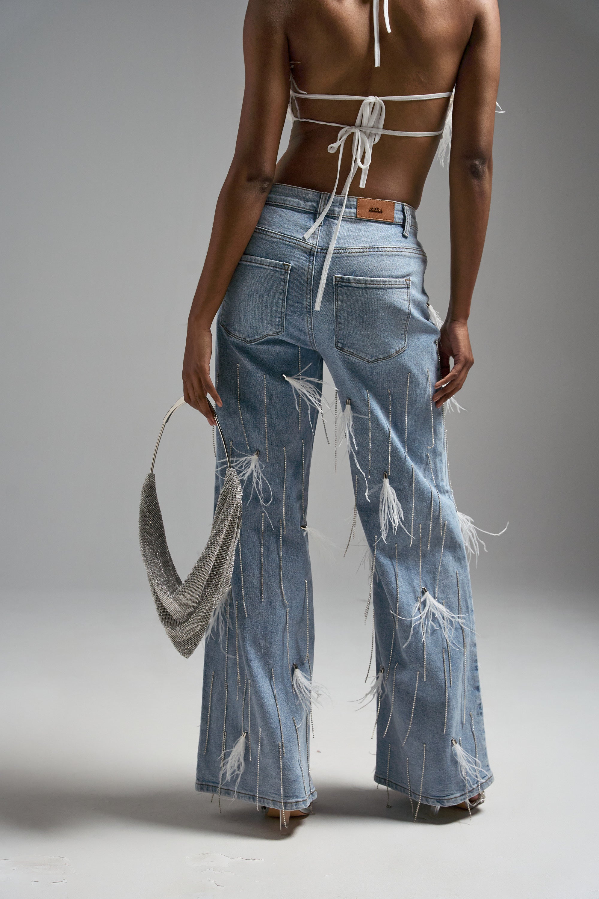 A person faces away in SHAKE IT FOR ME GIRL FEATHER EMBELLISHED DENIM with frayed strings, paired with a back-tie halter top. They hold a silver textured handbag in their left hand, set against a plain gray background.