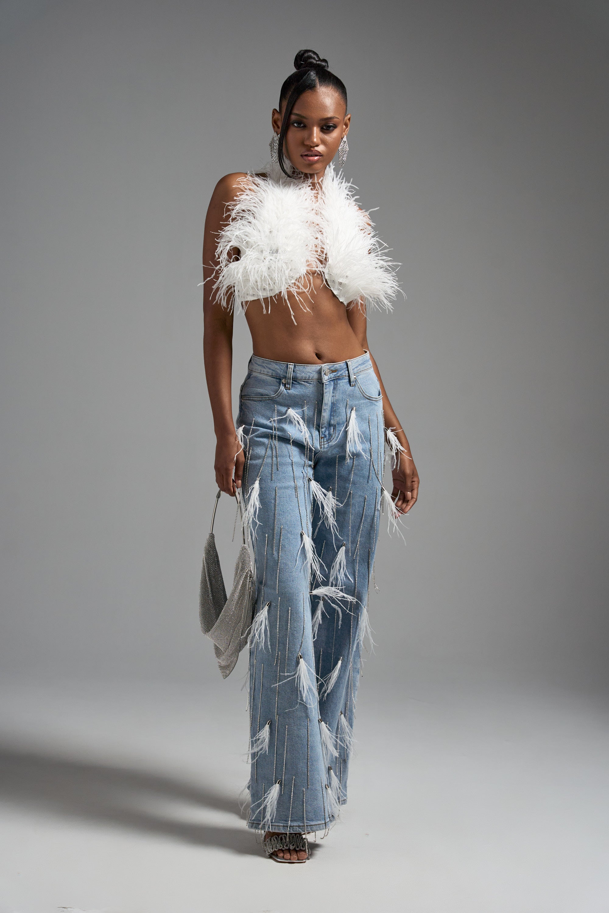 A woman poses confidently in the SHAKE IT FOR ME GIRL FEATHER EMBELLISHED DENIM, paired with a white feathered crop top and a small silver purse, standing against a plain gray background.