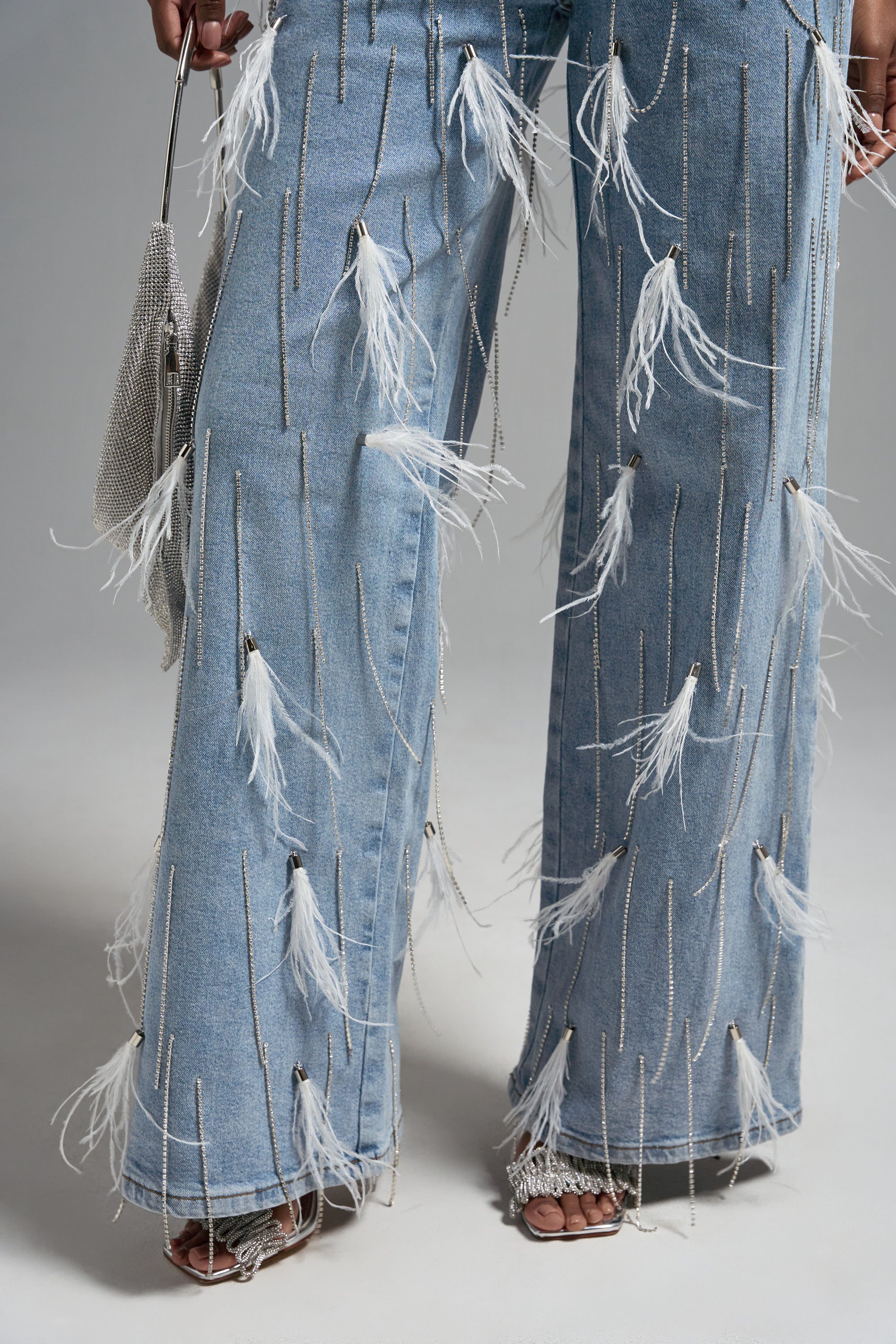 Close-up of a person wearing the SHAKE IT FOR ME GIRL FEATHER EMBELLISHED DENIM with dangling white feathers and silver chains, holding a sparkling silver purse and wearing open-toed heels.