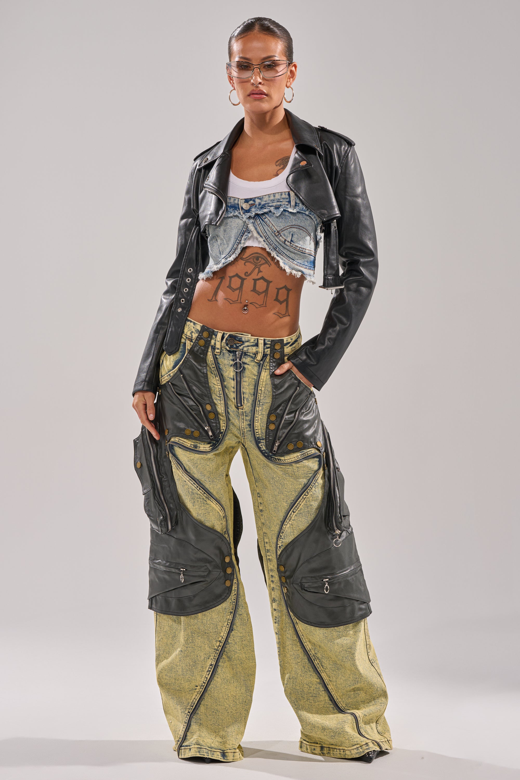 A person poses confidently in the CRAZY FOR YOU RELAXED DENIM, paired with a black leather jacket, distressed cropped denim top, hoop earrings, sunglasses, and midriff tattoos. 0