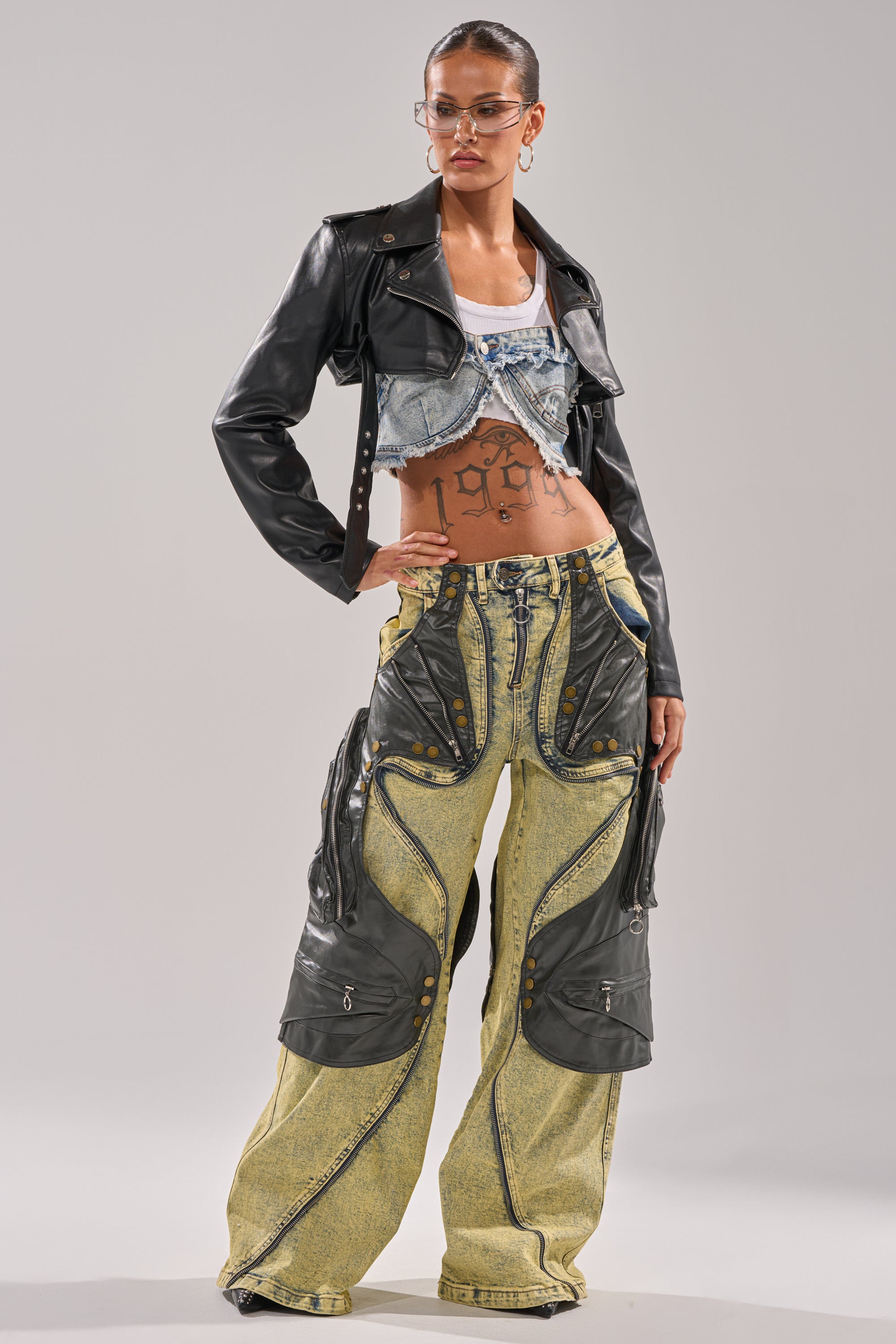 A woman poses confidently in the CRAZY FOR YOU RELAXED DENIM crop top, paired with a cropped black leather jacket and wide-leg yellow-green cargo pants, accessorized with glasses and hoop earrings against a plain background. 10