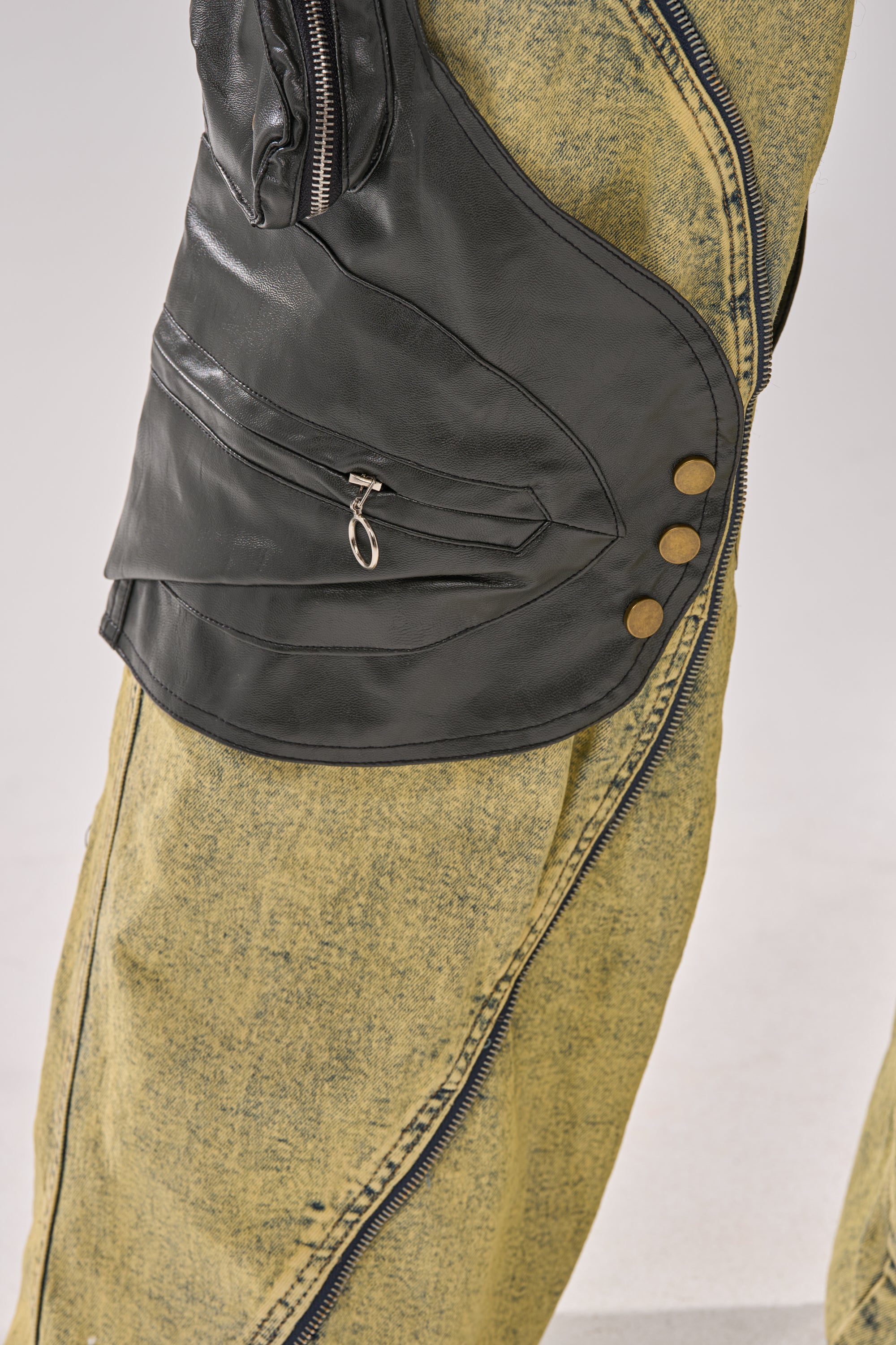 A close-up of a person wearing the CRAZY FOR YOU RELAXED DENIM in light yellow acid wash and a unique black vest with faux leather paneling, zippers, and metallic buttons. The scene is set against a plain light gray background. 12