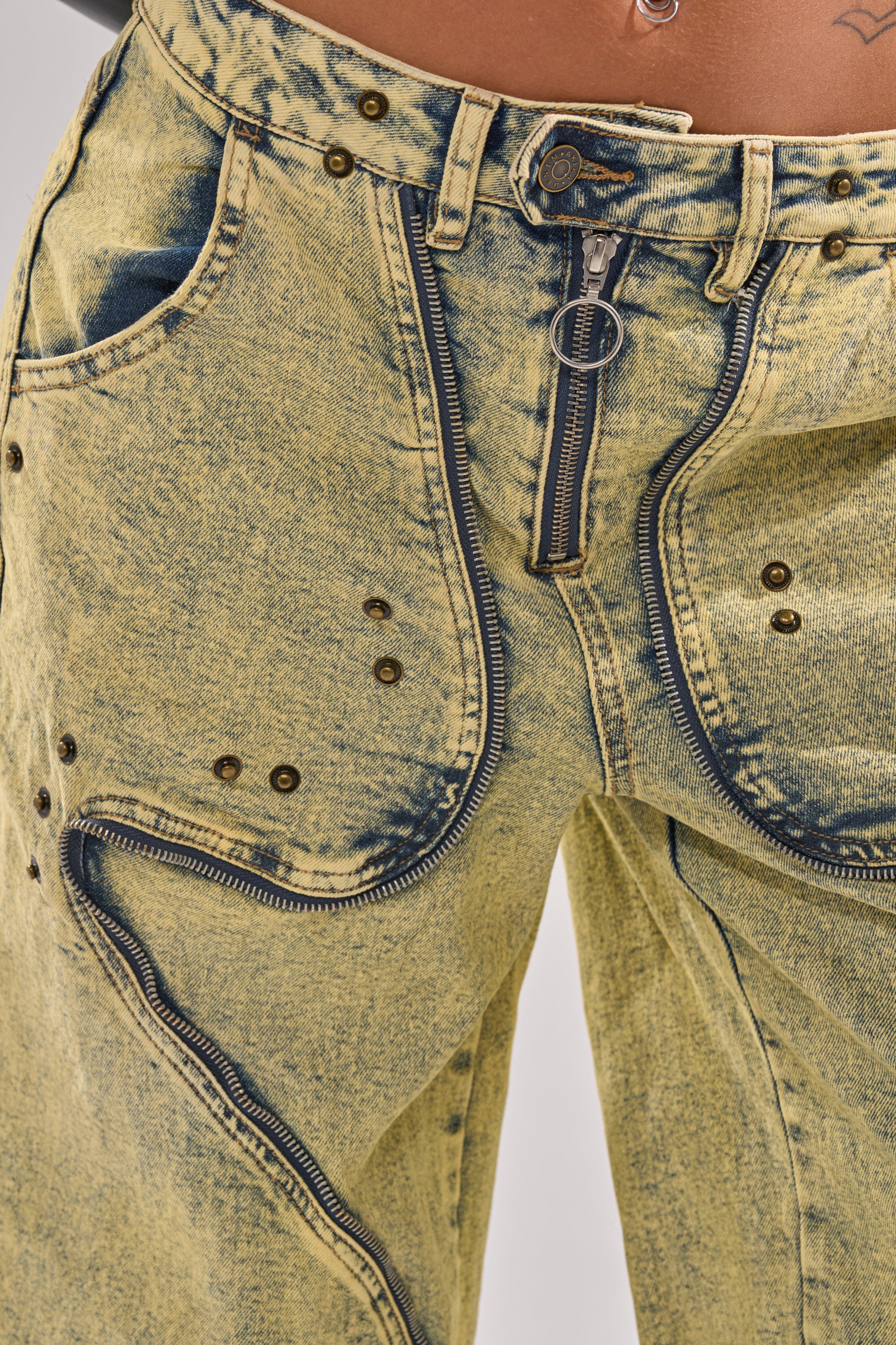 Close-up of a waist wearing CRAZY FOR YOU RELAXED DENIM in acid wash yellow with multiple zippers, metal rivets, and faded details; belly button piercing and tattoo above waistband capture the bold, relaxed vibe. 13