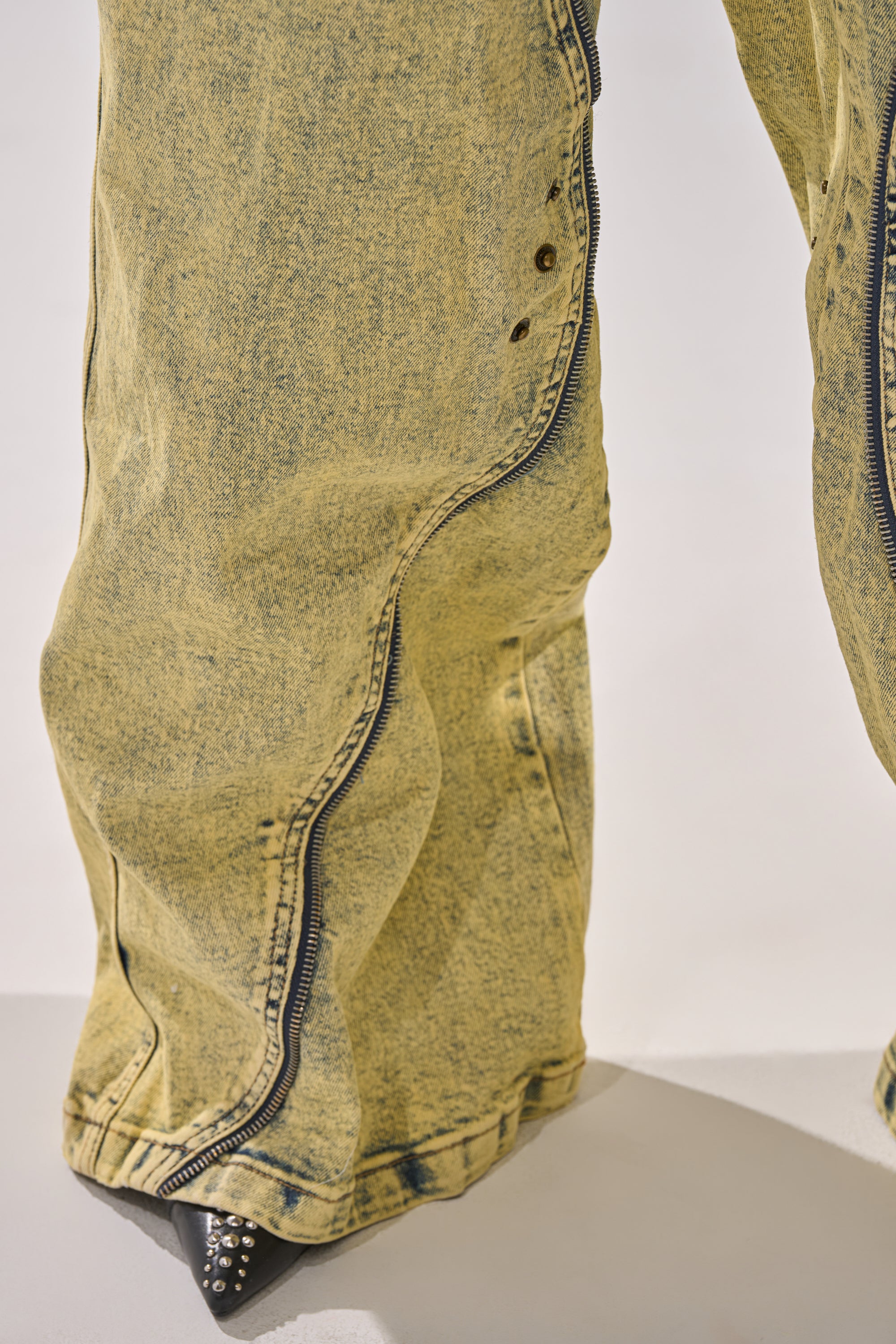 Close-up of CRAZY FOR YOU RELAXED DENIM in yellow acid wash with exposed zippers at the seams, styled with pointed black shoes featuring silver studs peeking from beneath the relaxed fit. 14