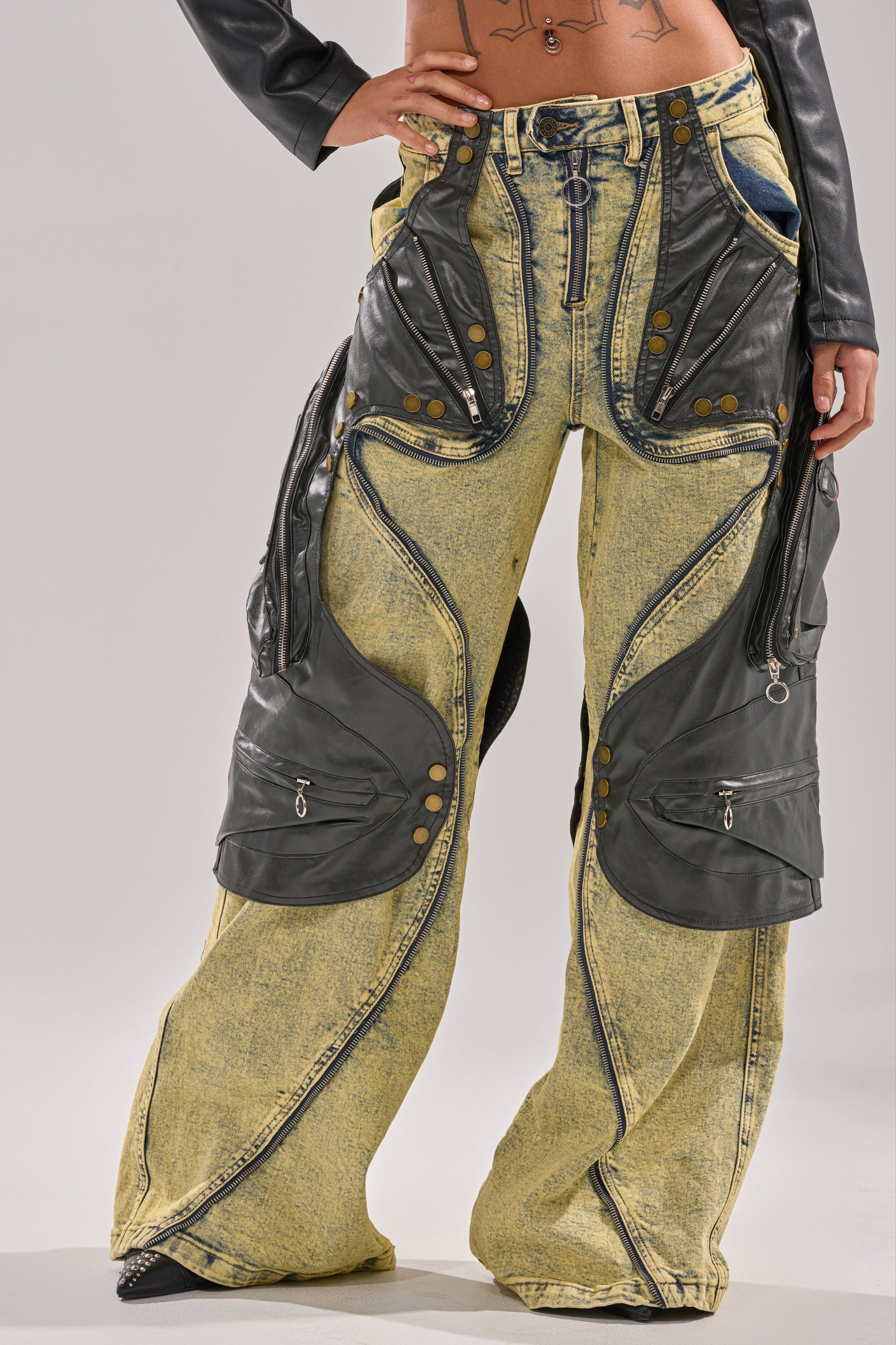 A person wearing CRAZY FOR YOU RELAXED DENIM—high-waisted, wide-leg yellow acid-wash jeans with faux leather panels, zippers, and metallic studs—poses with one hand on their hip and midriff exposed, showcasing tattoos. 3