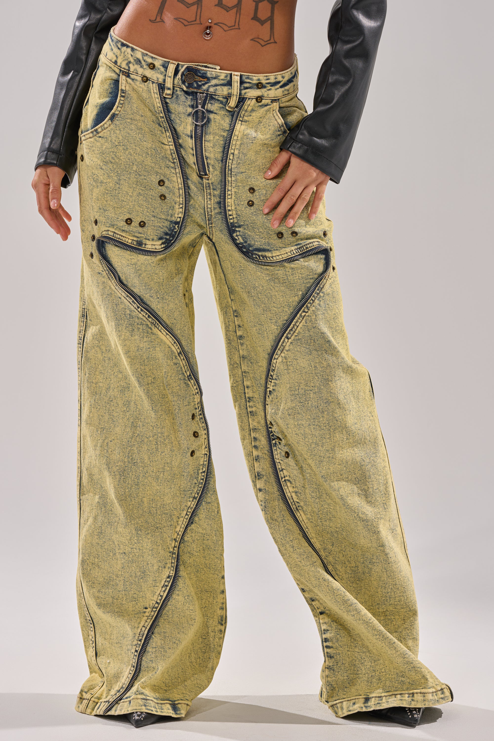 A person wears CRAZY FOR YOU RELAXED DENIM—acid wash jeans with dark seams, metal rivets, faux leather panels, and a lace-up waist—paired with a black crop top and black leather sleeves; only their lower torso and legs are shown. 4