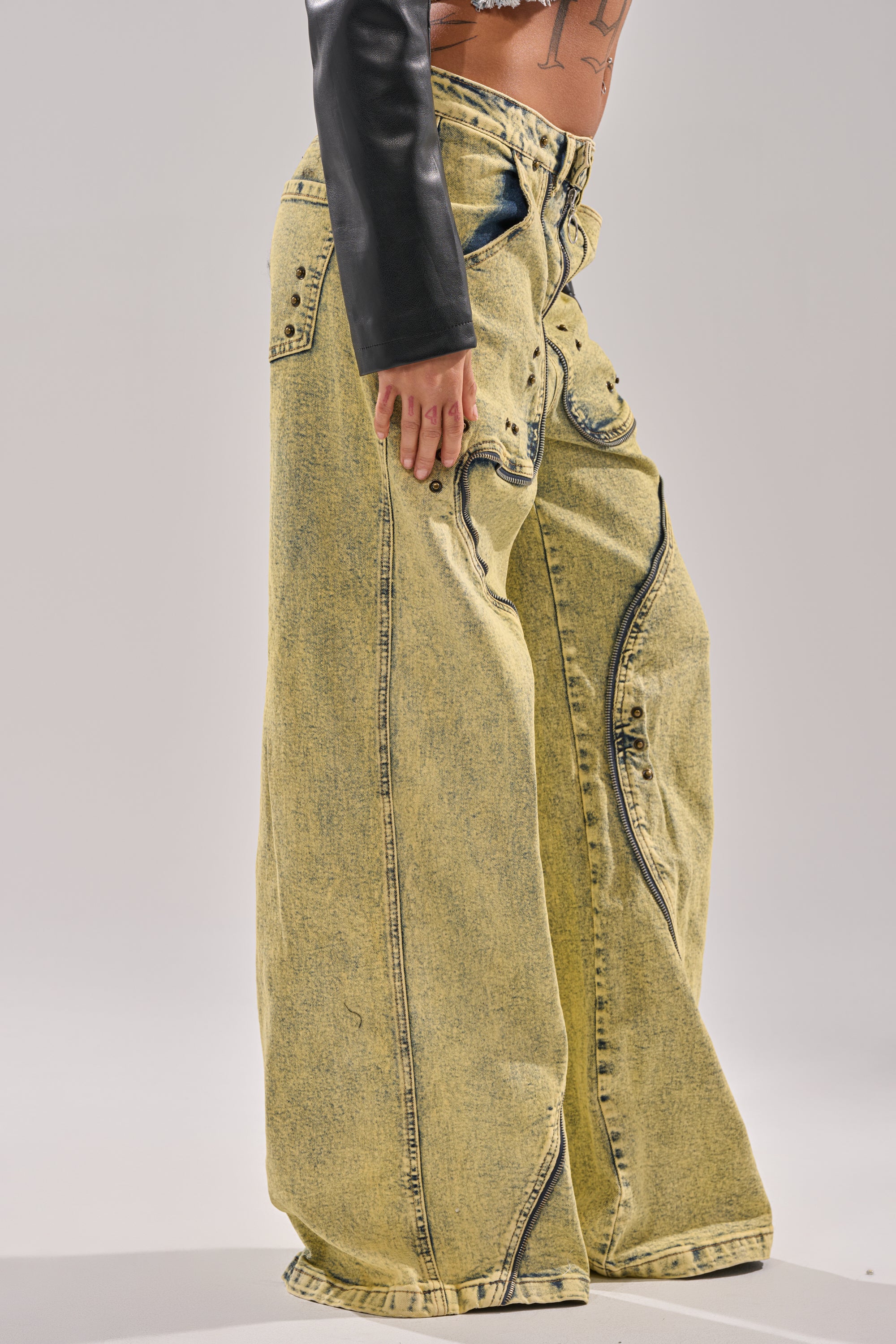 A person wearing CRAZY FOR YOU RELAXED DENIM—wide-leg yellow acid-wash jeans with faux leather panels and black rivet details. The image is cropped to show the lower torso and legs against a plain background. 6