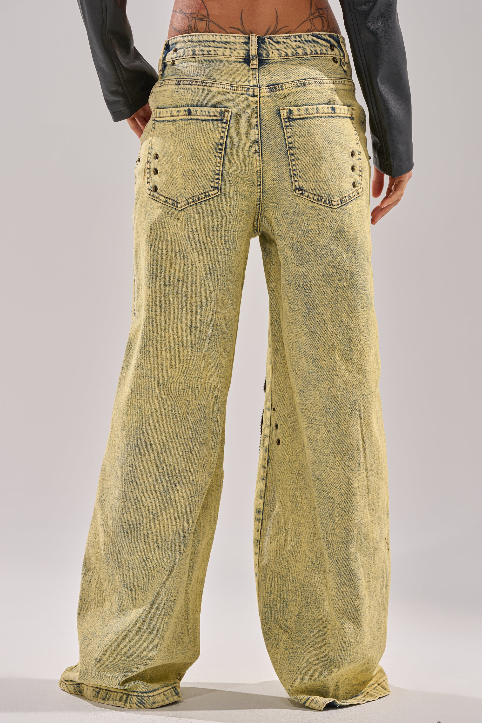 A person stands with their back to the camera, wearing CRAZY FOR YOU RELAXED DENIM acid-wash yellow jeans featuring two back pockets, decorative rivets, and a partially visible waistband and upper back. 8
