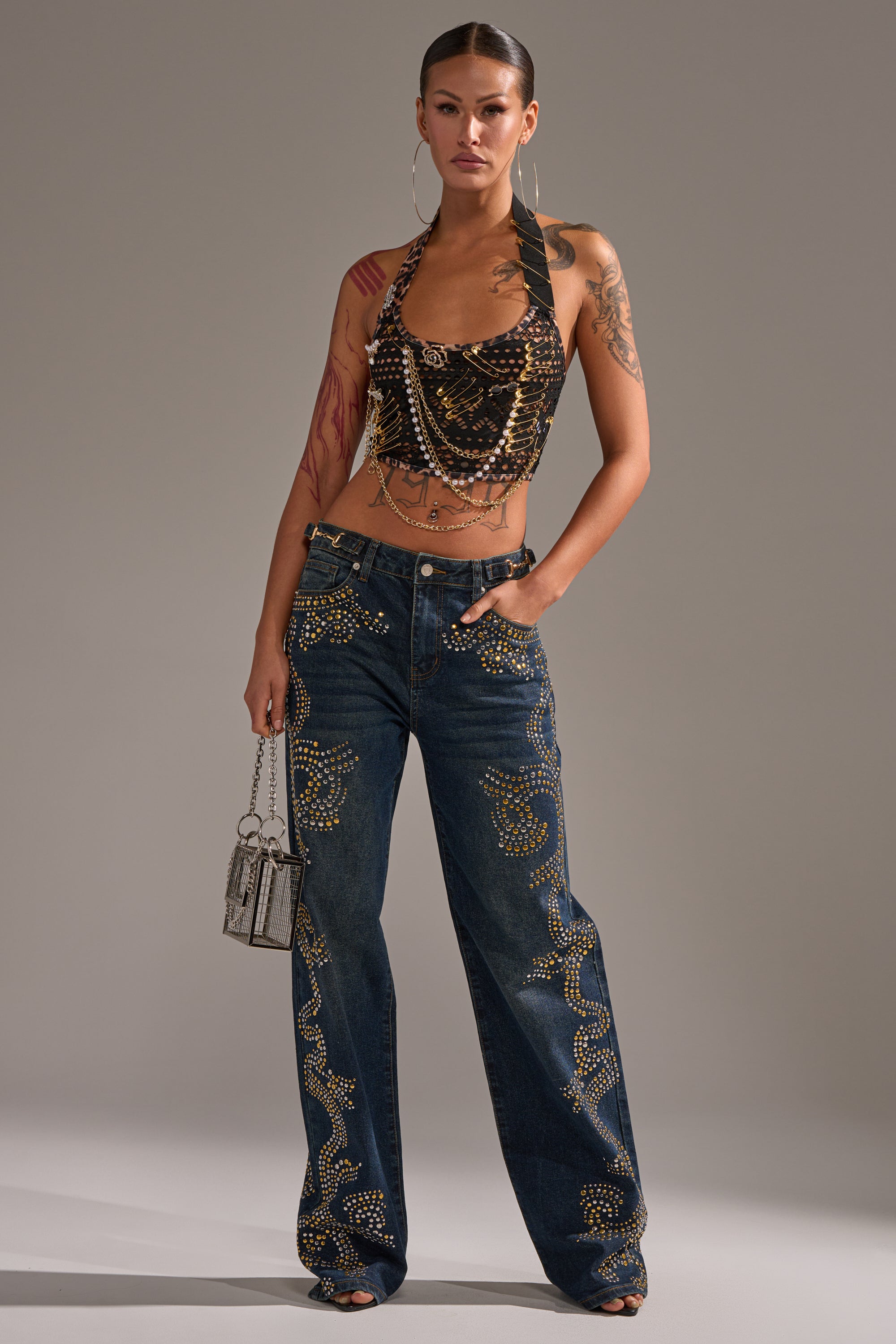 A woman with tattoos and pulled-back hair stands in a studio wearing ZIGGY STUDDED WIDE LEG DENIM, a black studded halter top, hoop earrings, and holding a small metallic handbag. 1