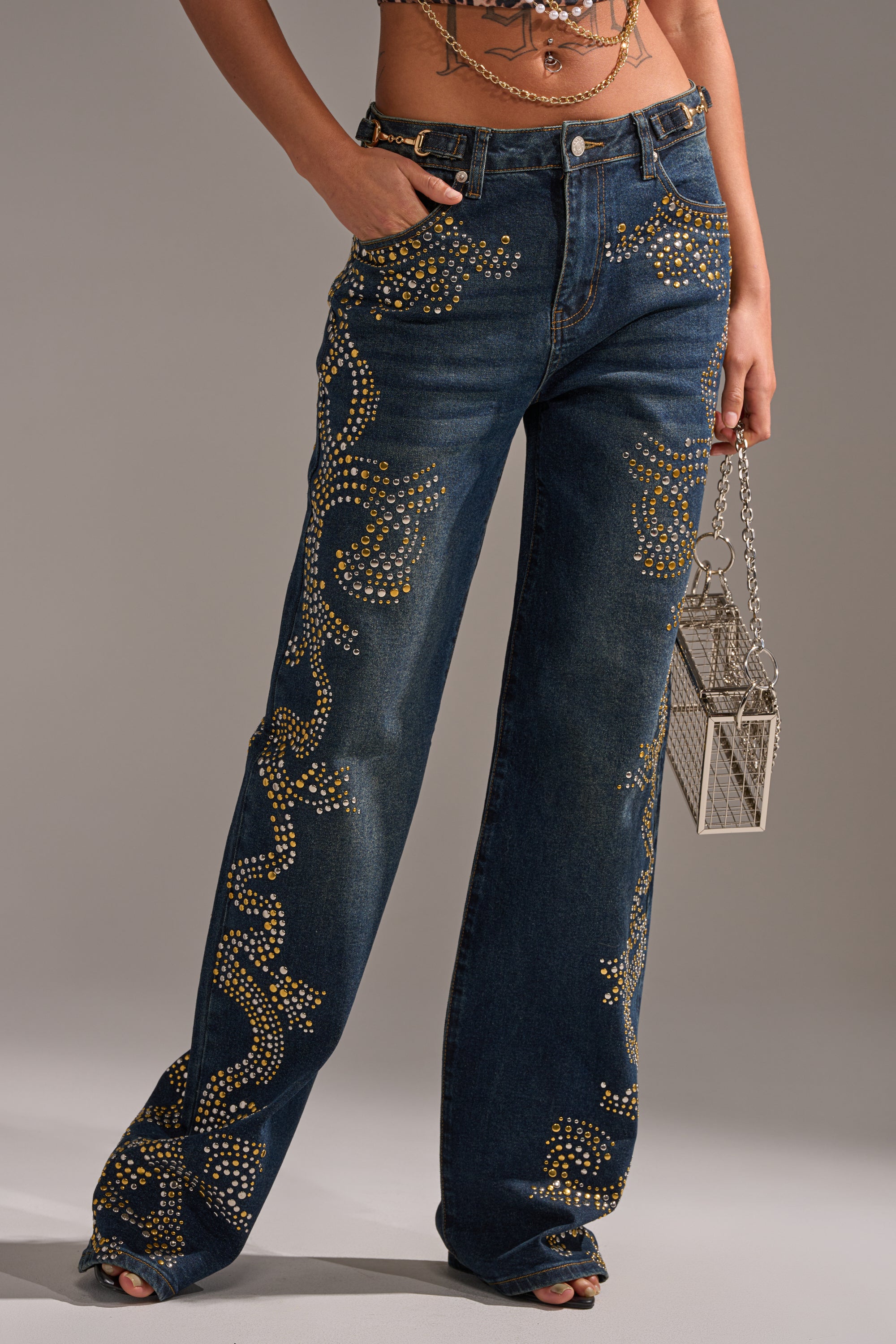 Wearing ZIGGY STUDDED WIDE LEG DENIM—high-rise, wide leg blue jeans with gold stud swirl designs—paired with a metallic box handbag and an ornate layered chain belt. 2