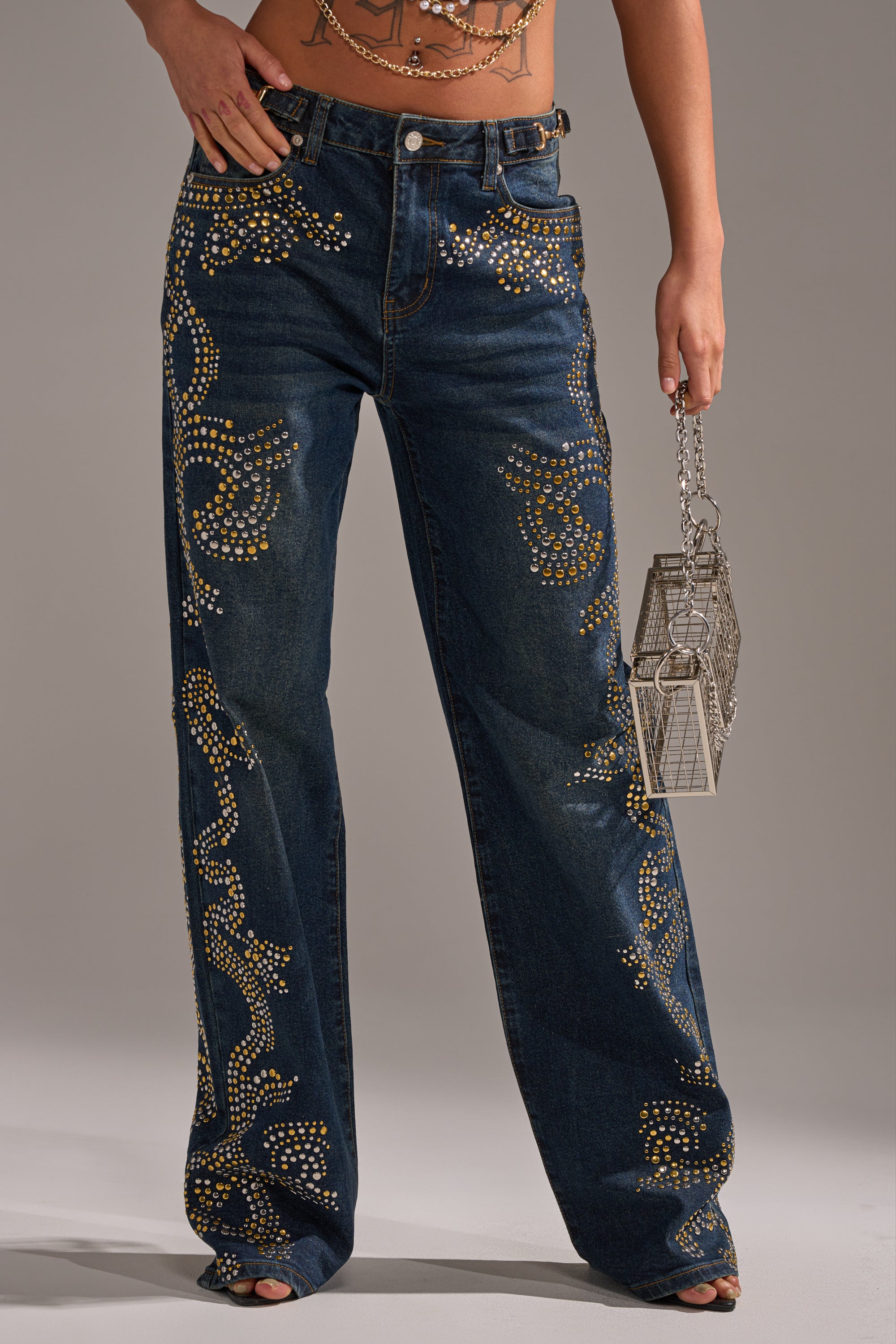 Wearing the ZIGGY STUDDED WIDE LEG DENIM, featuring high-rise, gold and silver swirl studs, a person holds a small metallic geometric handbag. Only the lower torso and legs are shown. 3
