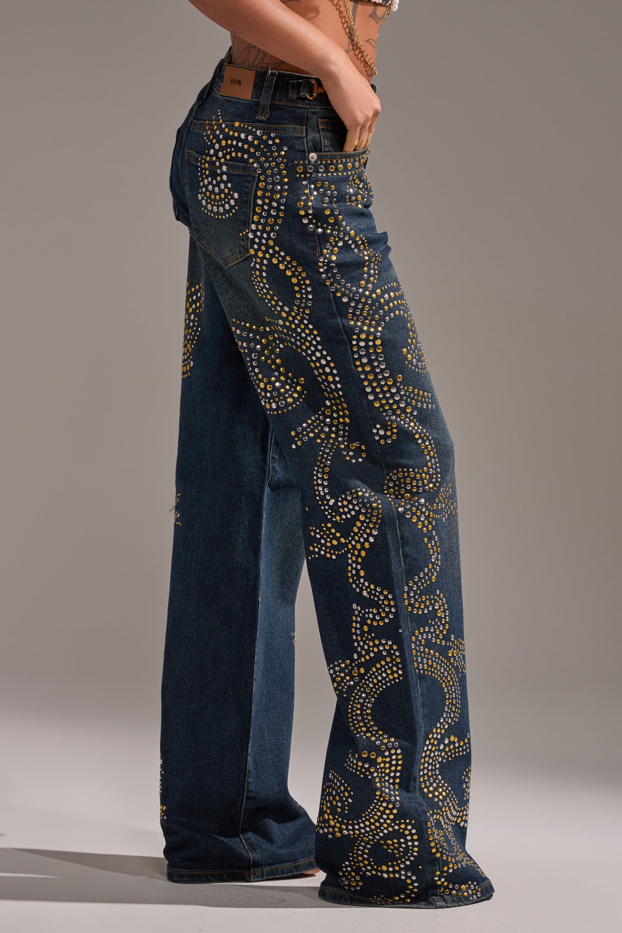 A person wears ZIGGY STUDDED WIDE LEG DENIM, blue jeans adorned with swirling gold studs, photographed from the waist down against a neutral background. 0
