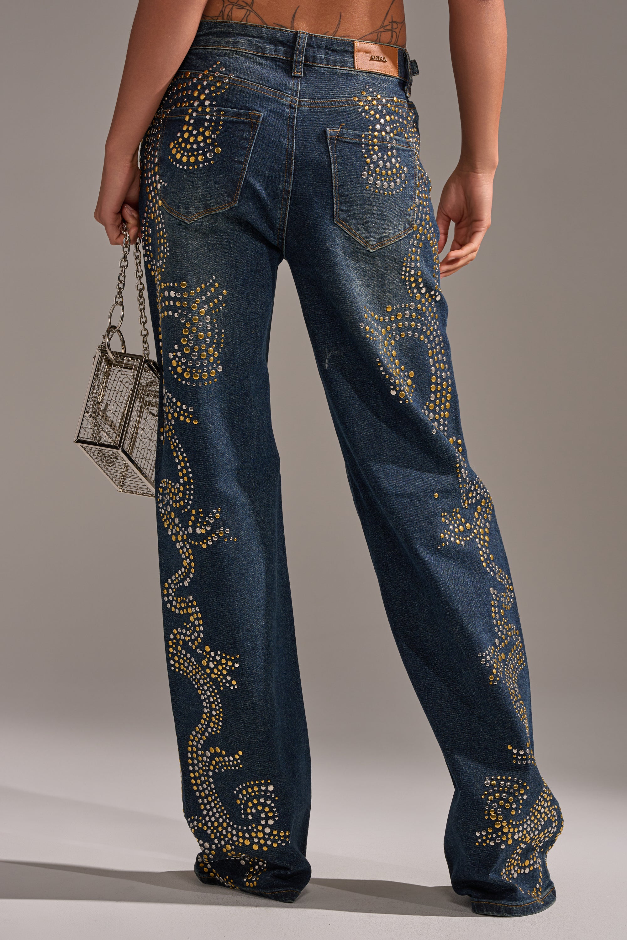 Someone stands with their back turned, wearing the ZIGGY STUDDED WIDE LEG DENIM—high-rise jeans featuring gold studded swirls. They carry a small silver handbag and display a tattoo on their lower back. 4