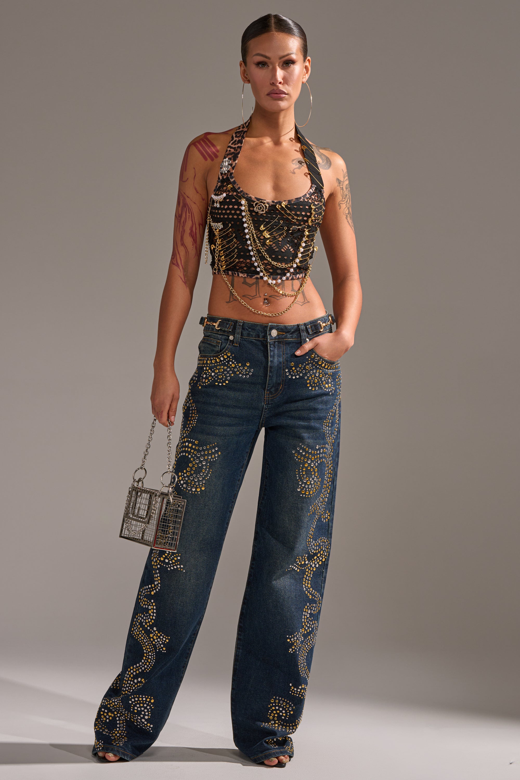 A woman exudes confidence in ZIGGY STUDDED WIDE LEG DENIM, paired with a beaded crop top and a metallic bag, her arm tattoos adding edge to the bold, gold-studded look. 5