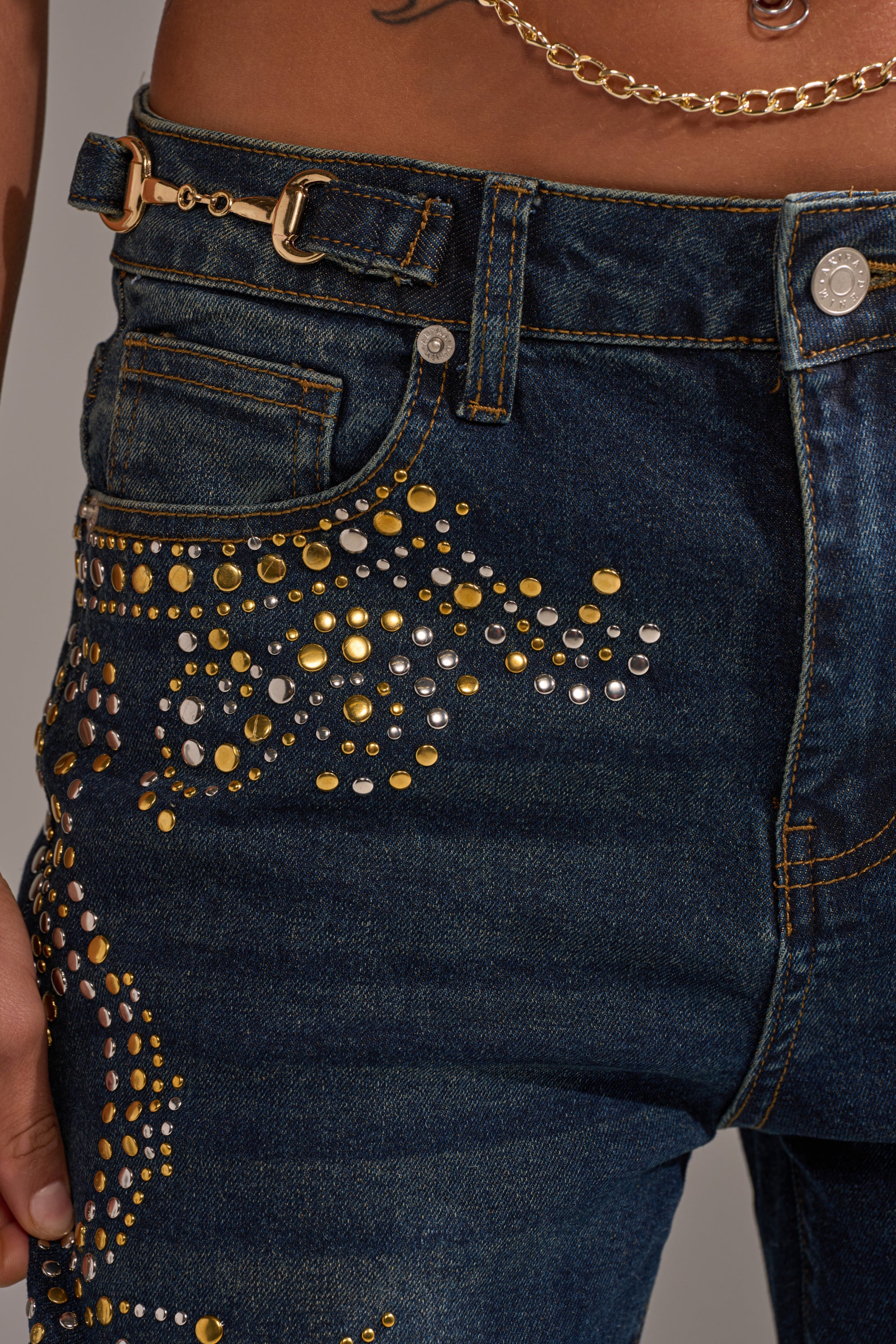 A close-up of the ZIGGY STUDDED WIDE LEG DENIM in dark blue, adorned with gold and silver studs in swirling patterns. A gold chain hangs from a belt loop, with part of a hand and waist visible. 6