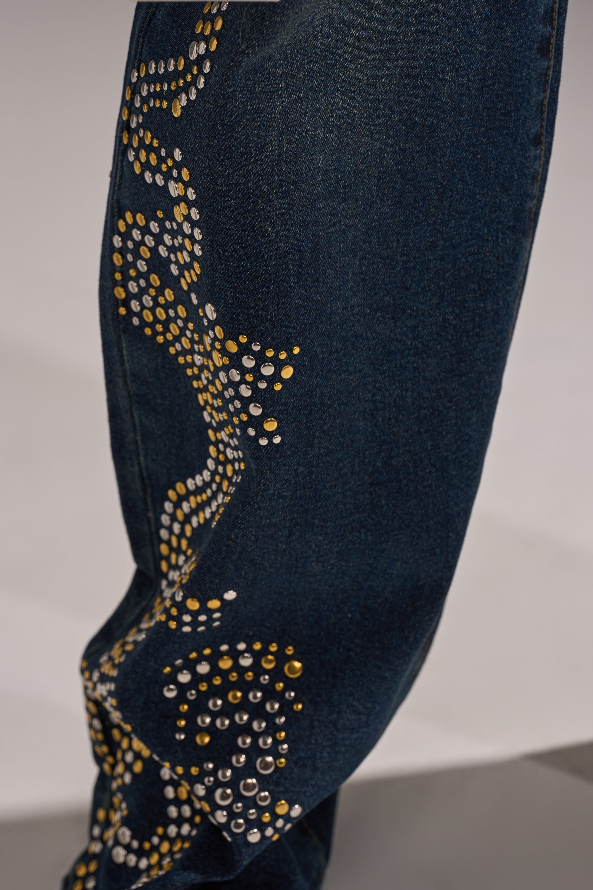 Close-up of the ZIGGY STUDDED WIDE LEG DENIM in dark blue, featuring a wide leg decorated with small silver and gold studs arranged in a curving pattern. 7