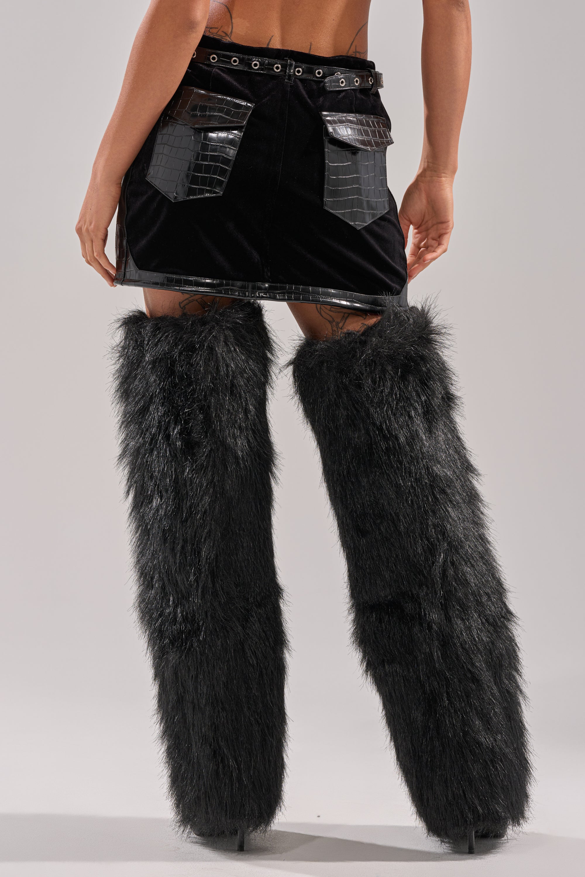 A person stands facing away, wearing the FEELING CRAZY MINI SKIRT in croc-embossed black faux leather with cargo pockets, paired with tall furry black leg warmers over high heels. The background is plain and light gray.