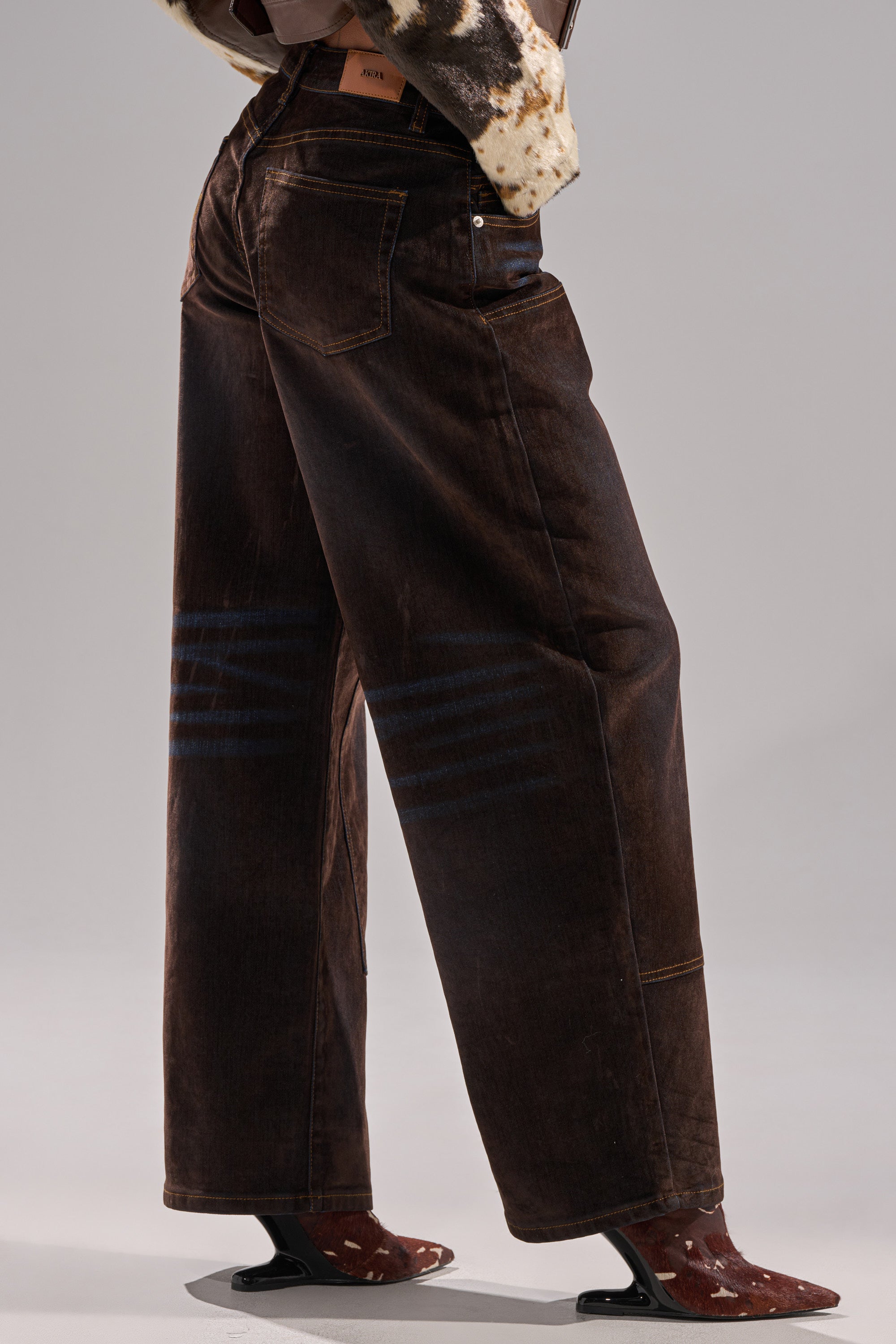 Wearing DAYLIGHT VELVET DENIM wide-leg, dark brown pants with blue stripes above the knees, paired with a cow-print jacket and high-heeled, see-through brown boots against a neutral gray background.