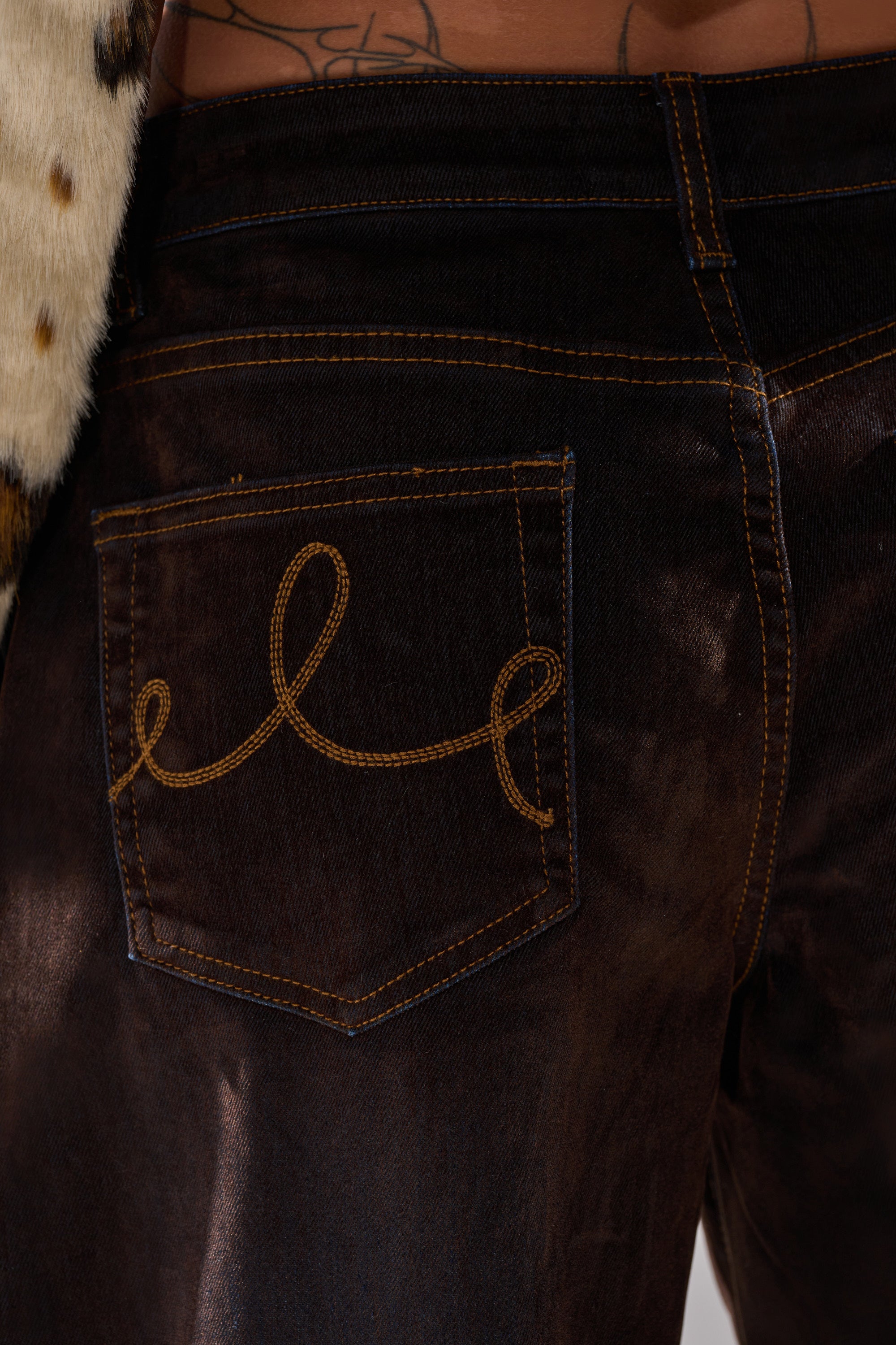 Close-up of the DAYLIGHT VELVET DENIM back pocket, featuring tan stitched looped designs. Visible are part of a tattooed lower back and a fur garment.