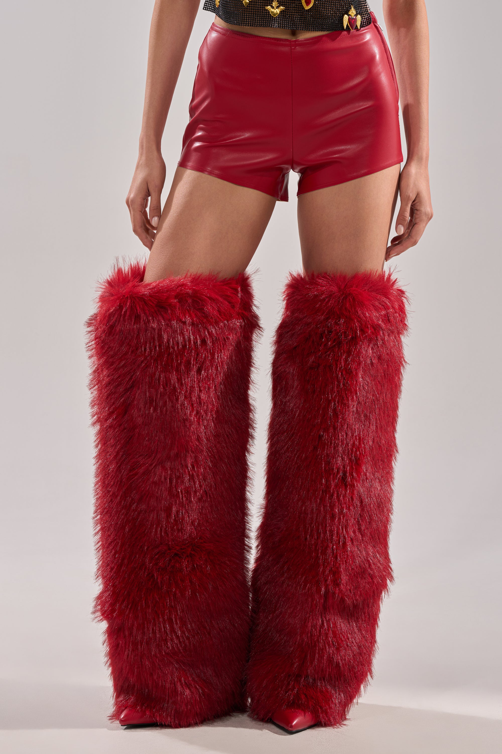 Someone wearing WONT LET IT GO FAUX LEATHER SHORT in shiny red vegan leather and tall, fluffy red furry leg warmers stands against a plain light background. 1