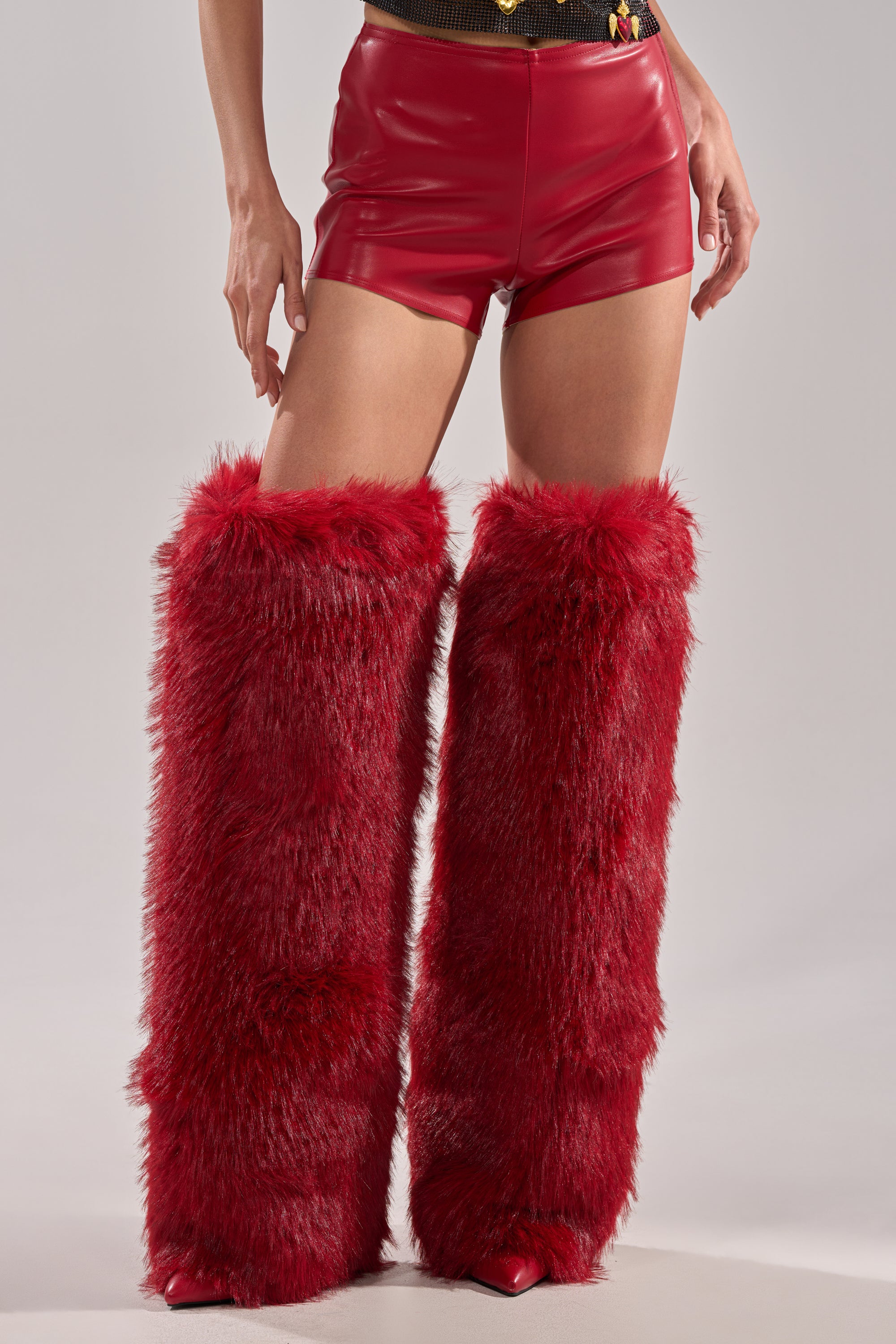 Wearing the WONT LET IT GO FAUX LEATHER SHORT in shiny red vegan leather, plus large fluffy red faux fur leg warmers and pointed red shoes, a person stands against a plain background. 2