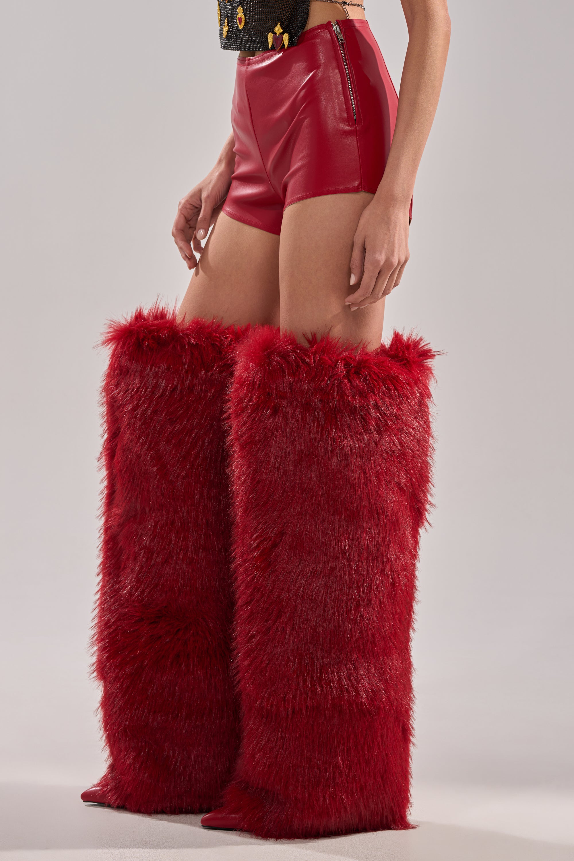 A person stands against a light background in red WONT LET IT GO FAUX LEATHER SHORTS, vibrant red faux fur knee-high boots, and a black top. The bold texture and color of the boots catch the eye. 3