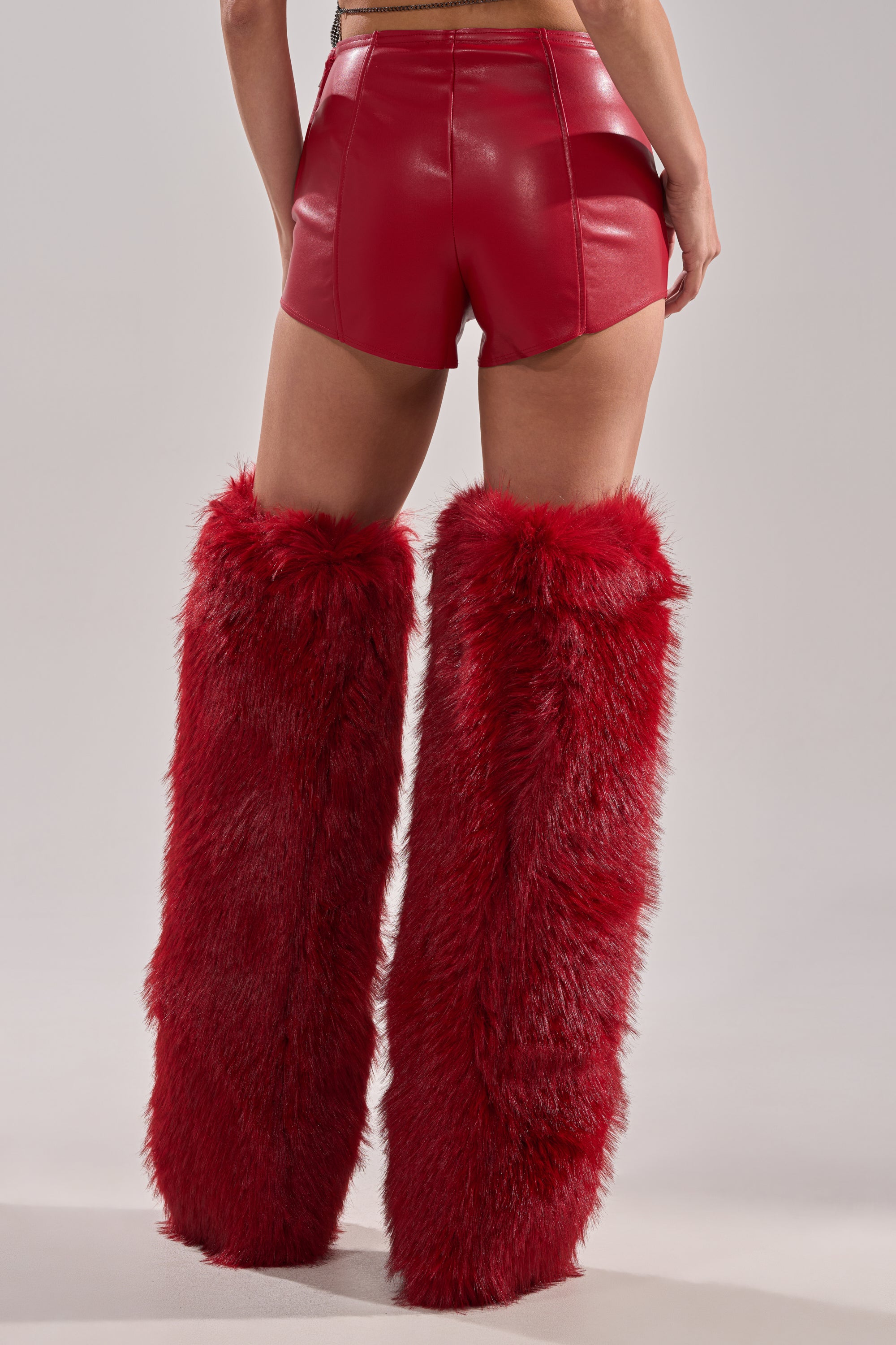A person wears WONT LET IT GO FAUX LEATHER SHORT in high-rise red vegan leather and tall, fluffy red faux fur boots, shown from the back against a plain light background. 4