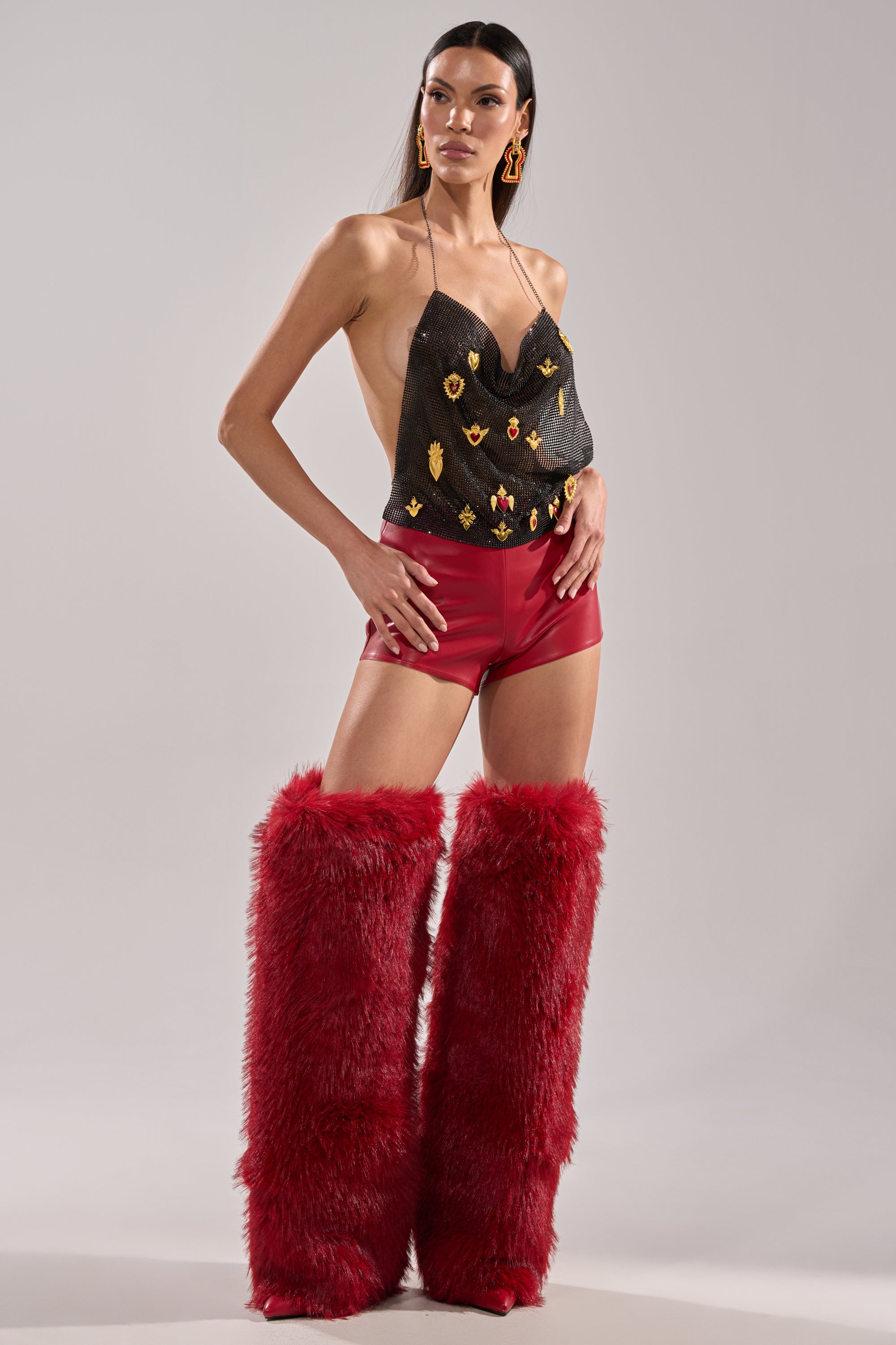 A woman poses in the WONT LET IT GO FAUX LEATHER SHORT, a black halter top with gold details, tall fluffy red over-the-knee boots, and bold gold earrings, standing against a plain backdrop. 5