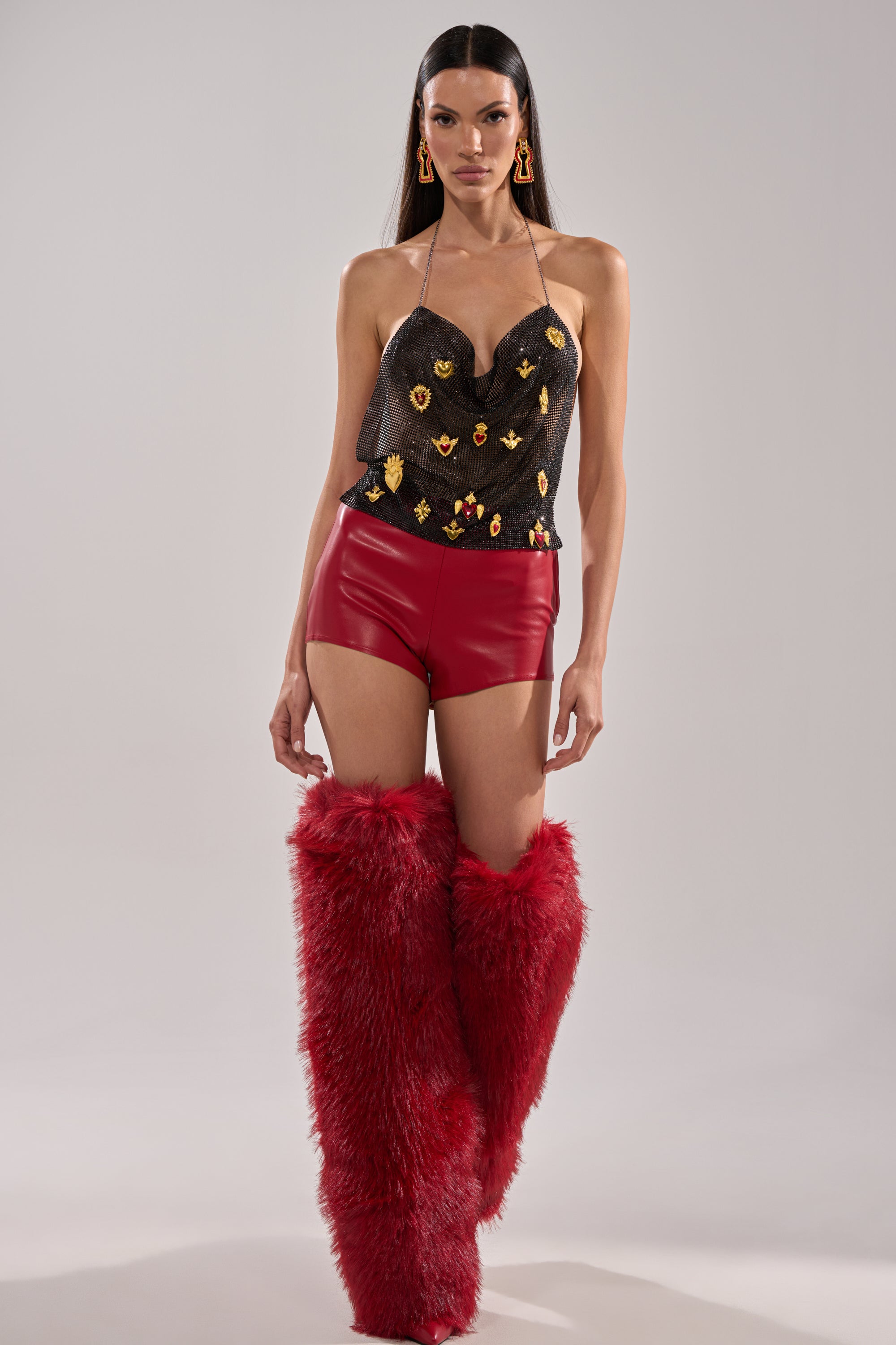 A woman stands confidently in WONT LET IT GO FAUX LEATHER SHORT—high-waisted red faux leather shorts—paired with a black sequined halter top, tall red fur boots, statement earrings, and long straight dark hair. 6