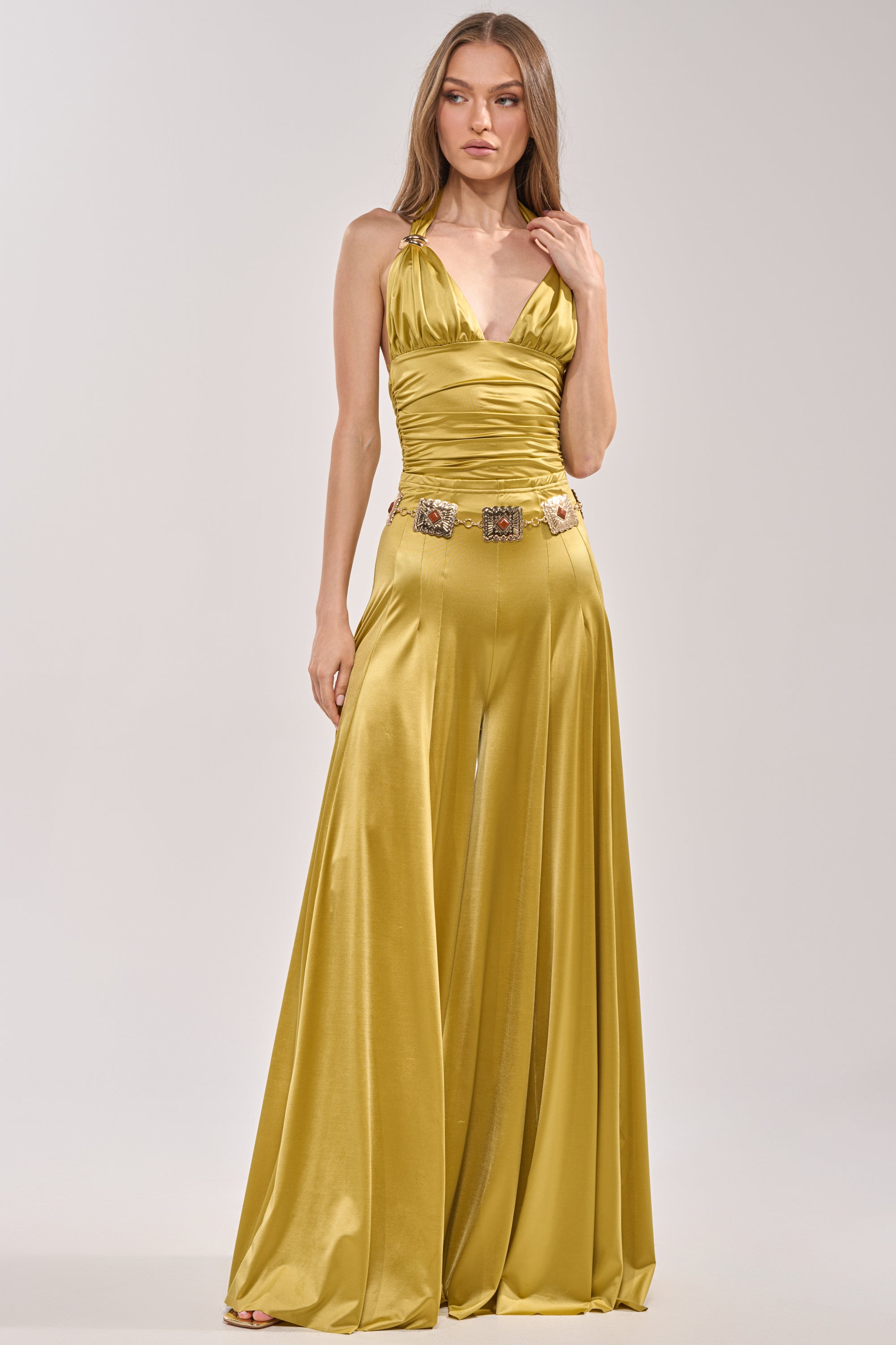 A woman in the DANCE THE NIGHT AWAY SLINKY TOP—a sleeveless, ruched halter gold satin jumpsuit with wide-leg pants and a jeweled belt—stands against a plain light background. 6