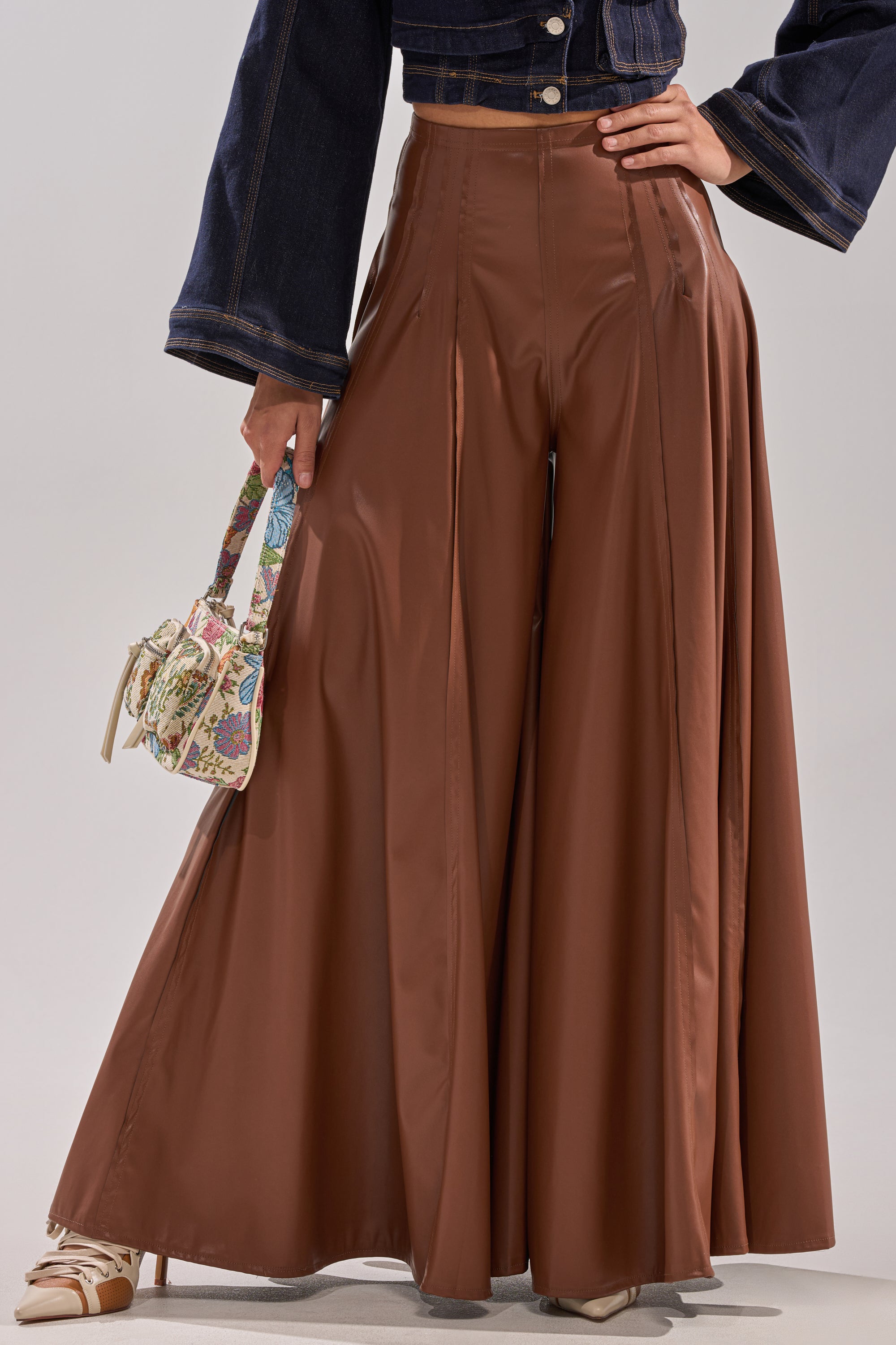 A person wearing GALAXY FAUX LEATHER TROUSER IN BROWN, a cropped dark denim jacket, cream heels, and holding a small floral handbag. The image is cropped at the shoulders. 0