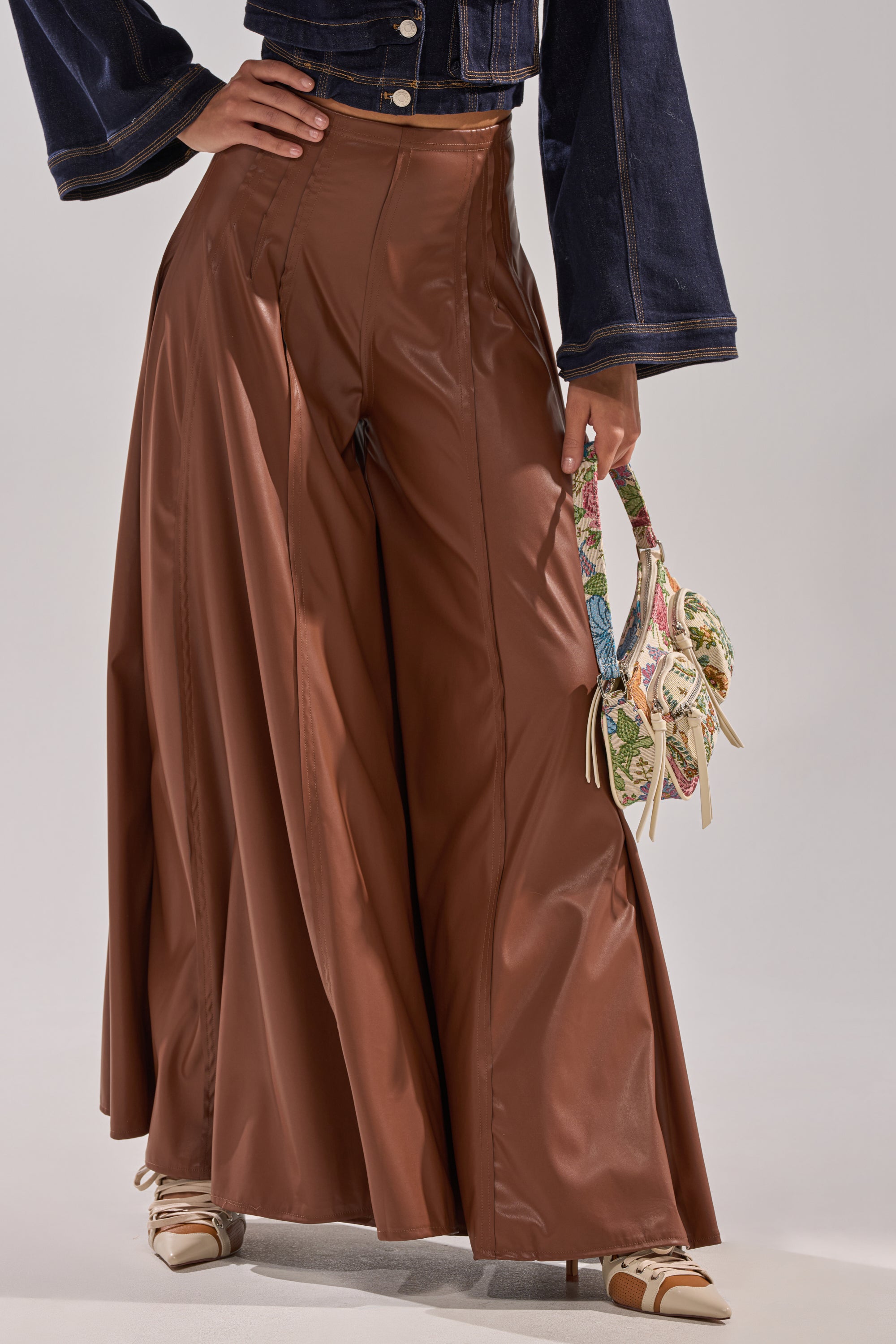 A person wears the GALAXY FAUX LEATHER TROUSER IN BROWN, paired with a dark denim jacket, pointed-toe shoes, and holds a small floral handbag. The photo is cropped at the waist. 1