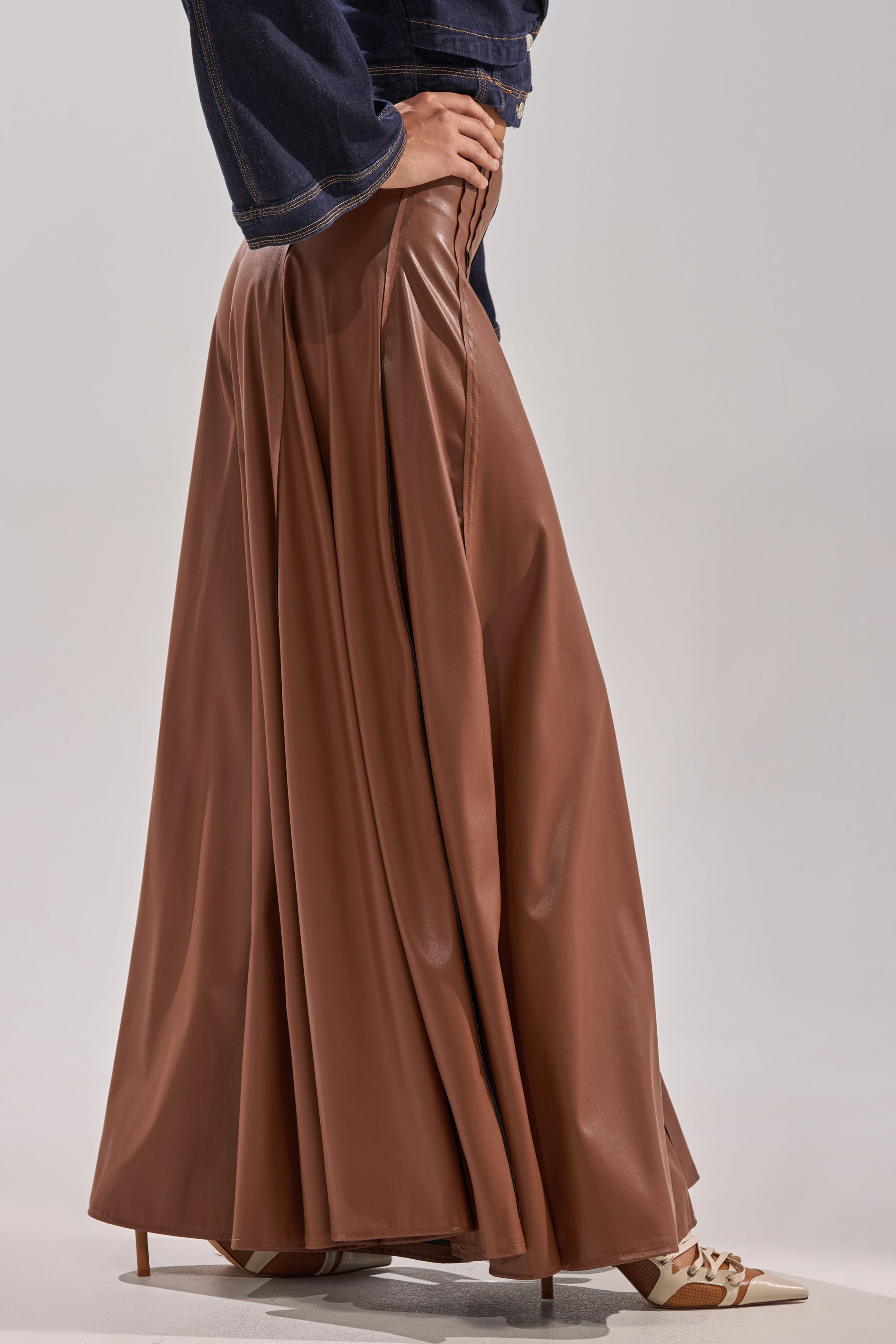 Wearing the GALAXY FAUX LEATHER TROUSER IN BROWN, tan pointed-toe heels, and a dark denim jacket, a person stands against a plain light background. 2