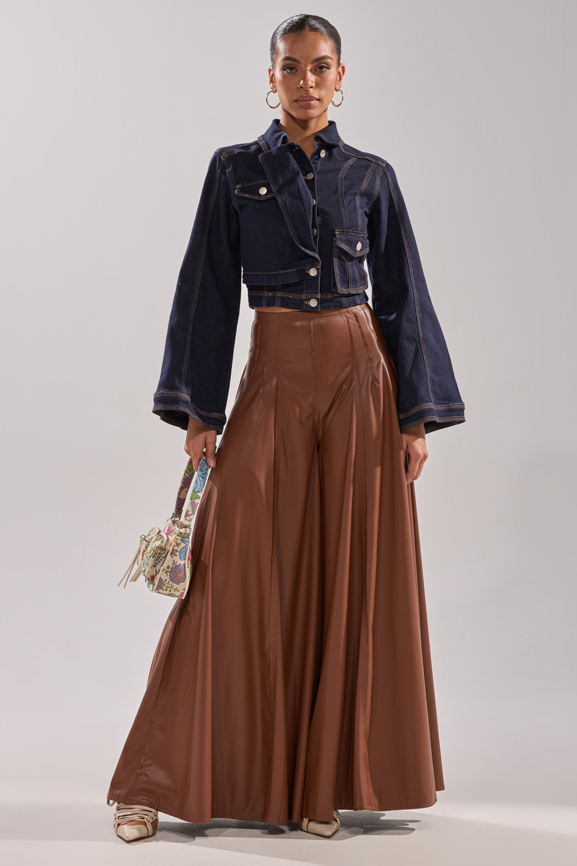 A woman wears high-waisted GALAXY FAUX LEATHER TROUSER IN BROWN, a dark denim cropped jacket, beige pointed shoes, and holds a small patterned handbag. Her hair is tied back and she accessorizes with hoop earrings. 4