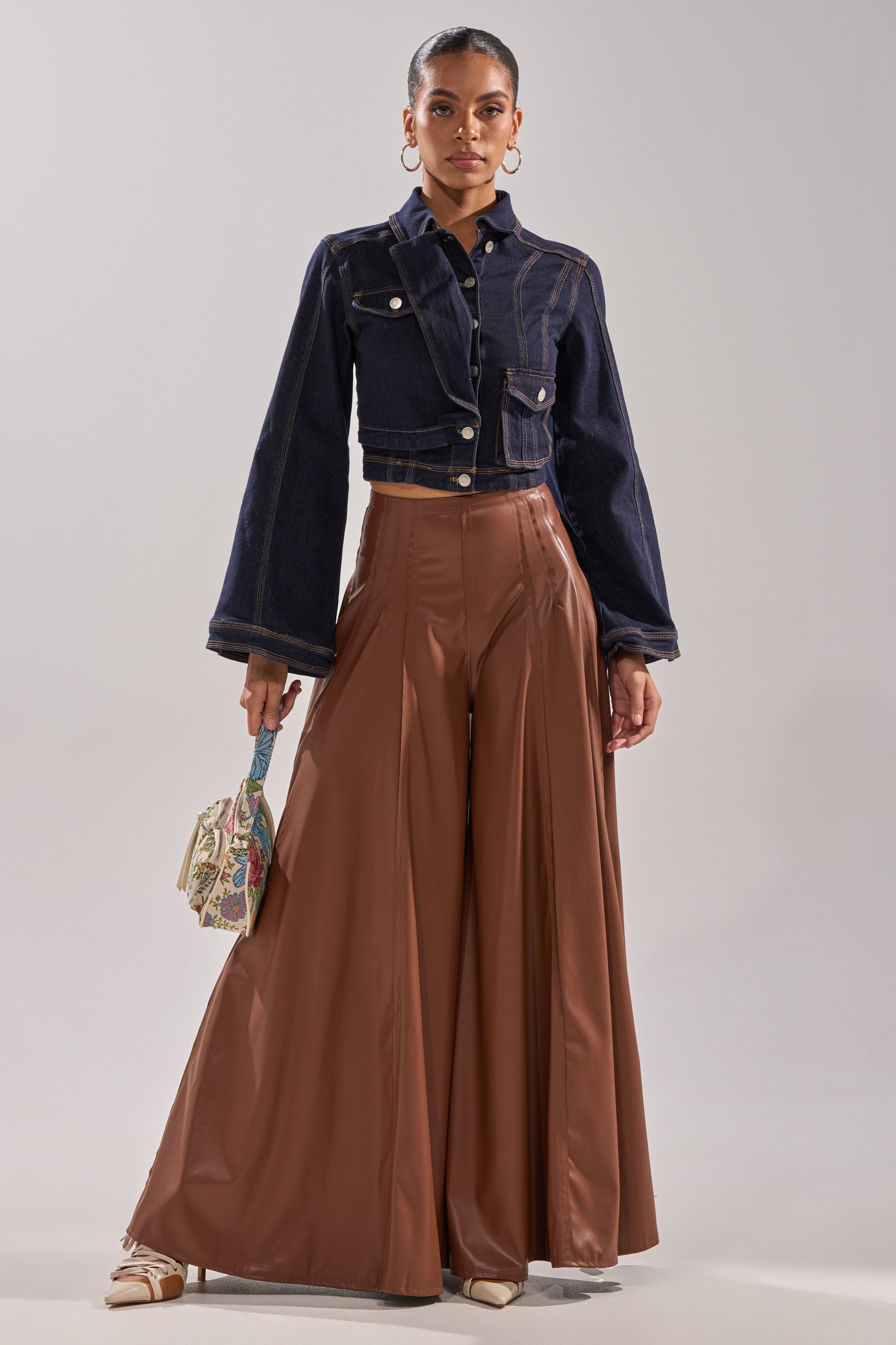 A woman stands confidently in a cropped dark denim jacket, GALAXY FAUX LEATHER TROUSER IN BROWN, beige heels, and holds a small decorative handbag. She accessorizes with hoop earrings and wears her hair tied back. 7