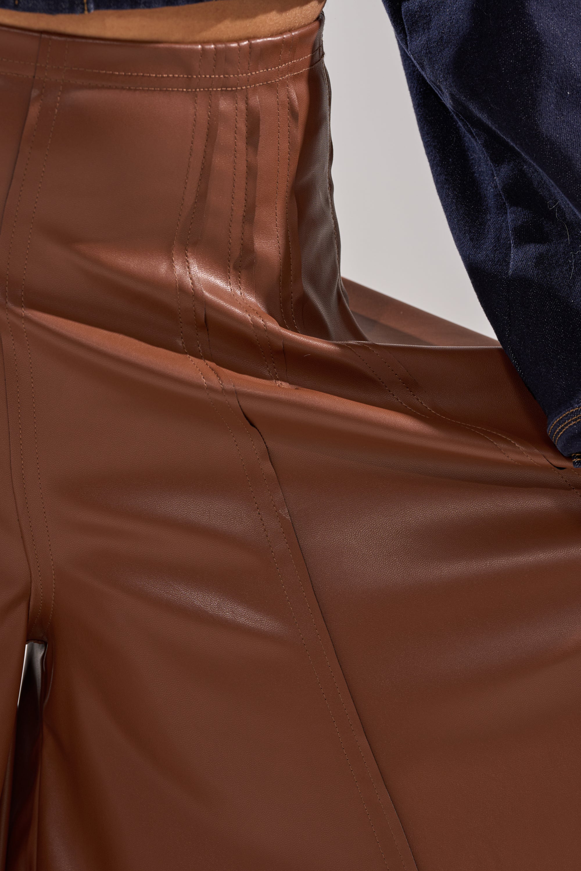 Close-up of someone wearing the GALAXY FAUX LEATHER TROUSER IN BROWN—high-waisted, shiny vegan leather pants with prominent stitching and a glossy texture—paired with a dark blue long-sleeve top for an eco-friendly classic style. 8