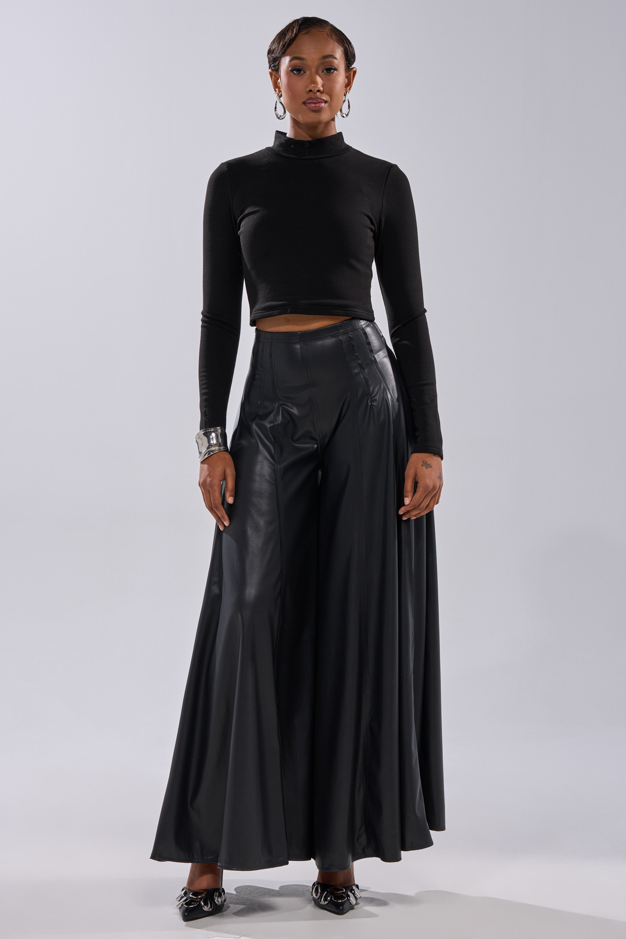 A person stands against a plain background wearing a black long-sleeve crop top, GALAXY FAUX LEATHER TROUSER, black pointed shoes, hoop earrings, and a silver cuff bracelet. 0