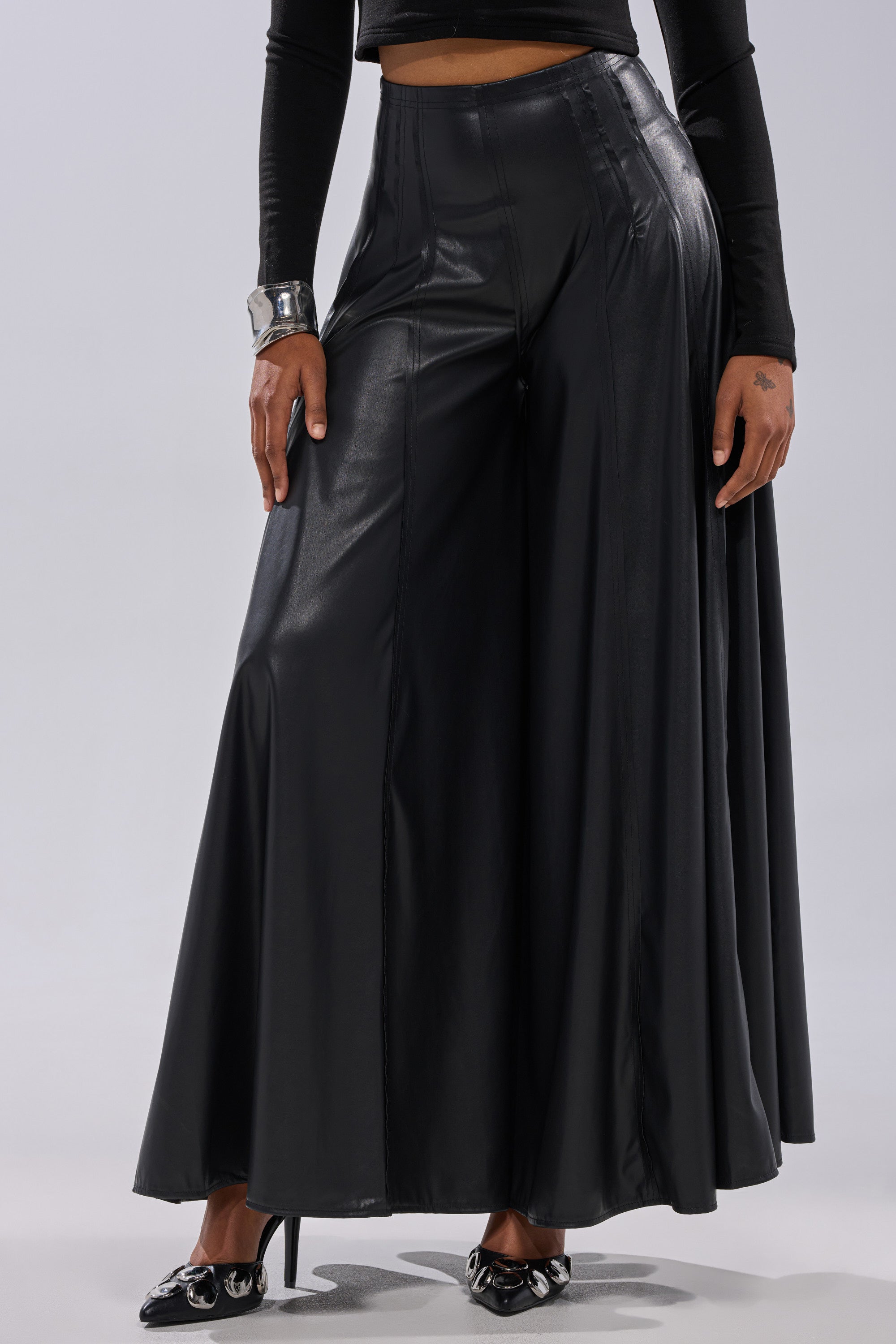 Wearing the GALAXY FAUX LEATHER TROUSER, a long-sleeve black crop top, shiny black heels, and a chunky silver bracelet, a person stands against a plain light background. 1