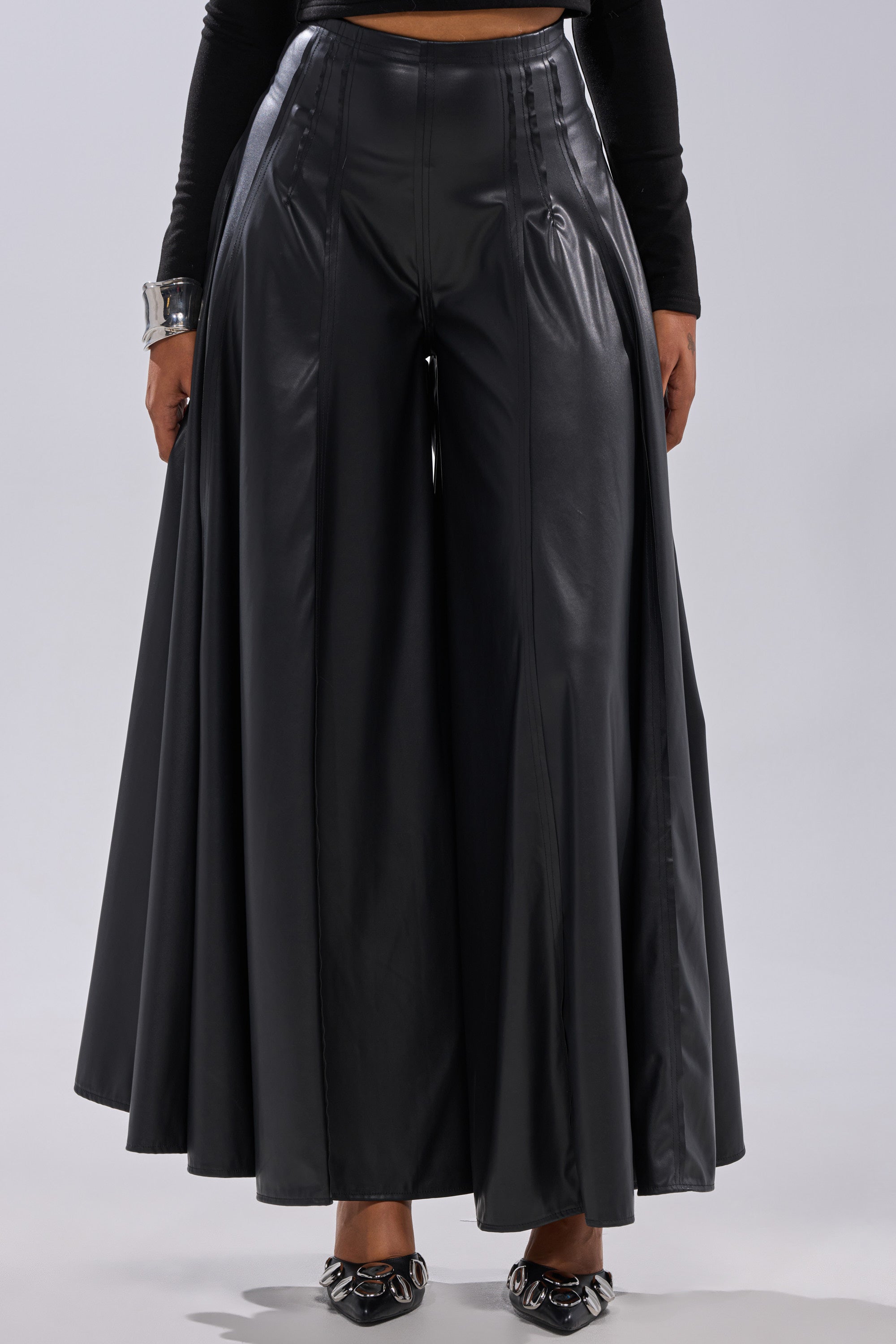 A person wears GALAXY FAUX LEATHER TROUSER—high-waisted, wide-leg black faux leather pants—styled with a black long-sleeve top, silver bracelet, and pointed black shoes. 2