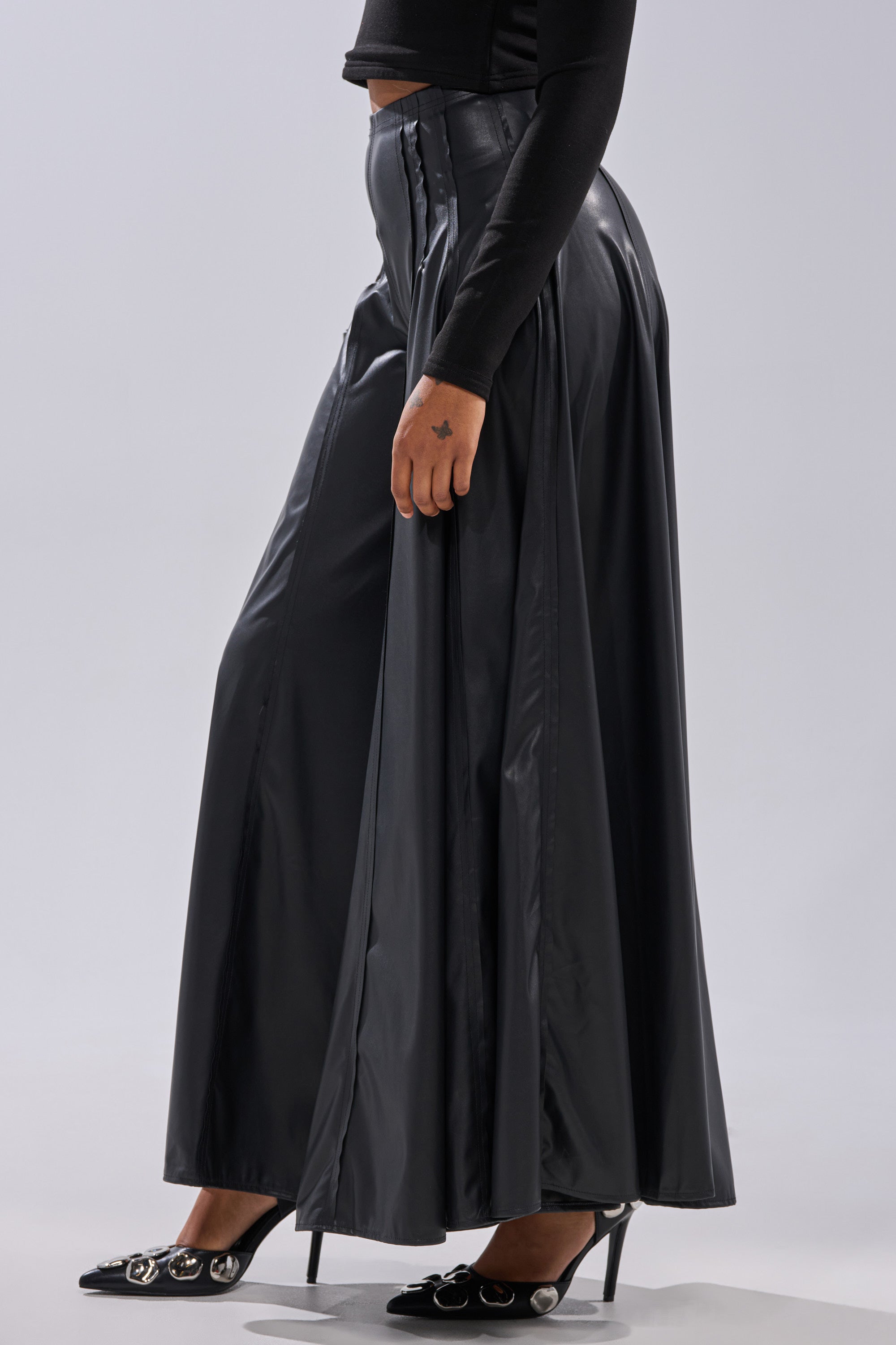 A person wears the GALAXY FAUX LEATHER TROUSER with a long-sleeve black top and black pointed high-heeled shoes featuring decorative buckles, standing against a plain background. 3