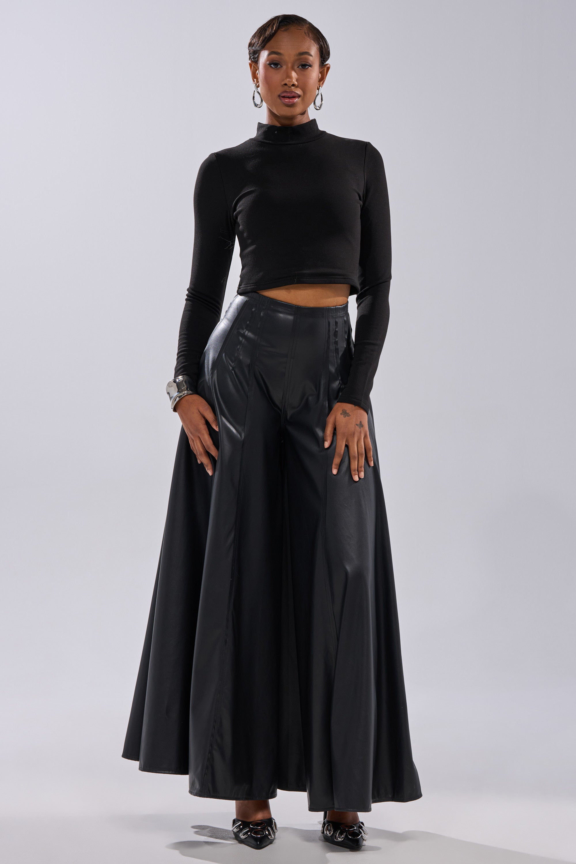 A woman poses confidently in the GALAXY FAUX LEATHER TROUSER, featuring a high-rise waistband and wide legs, paired with a black long-sleeve crop top, hoop earrings, bracelets, and black pointed shoes against a light background. 5