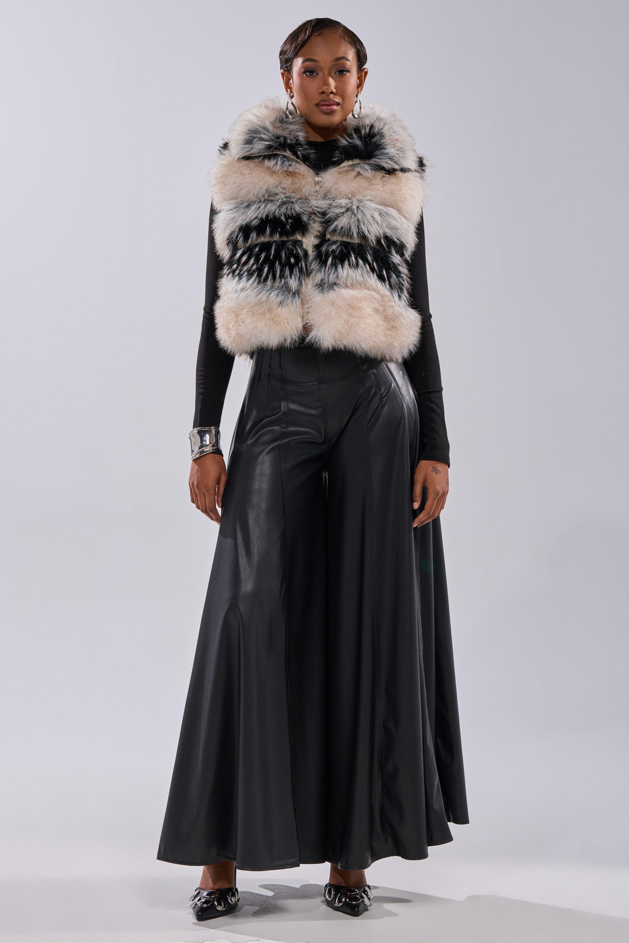 A woman stands facing forward, wearing a black long-sleeve top, a patterned faux fur vest, the GALAXY FAUX LEATHER TROUSER with a high-rise waist and wide legs, black pointed-toe shoes, and a chunky bracelet. 6