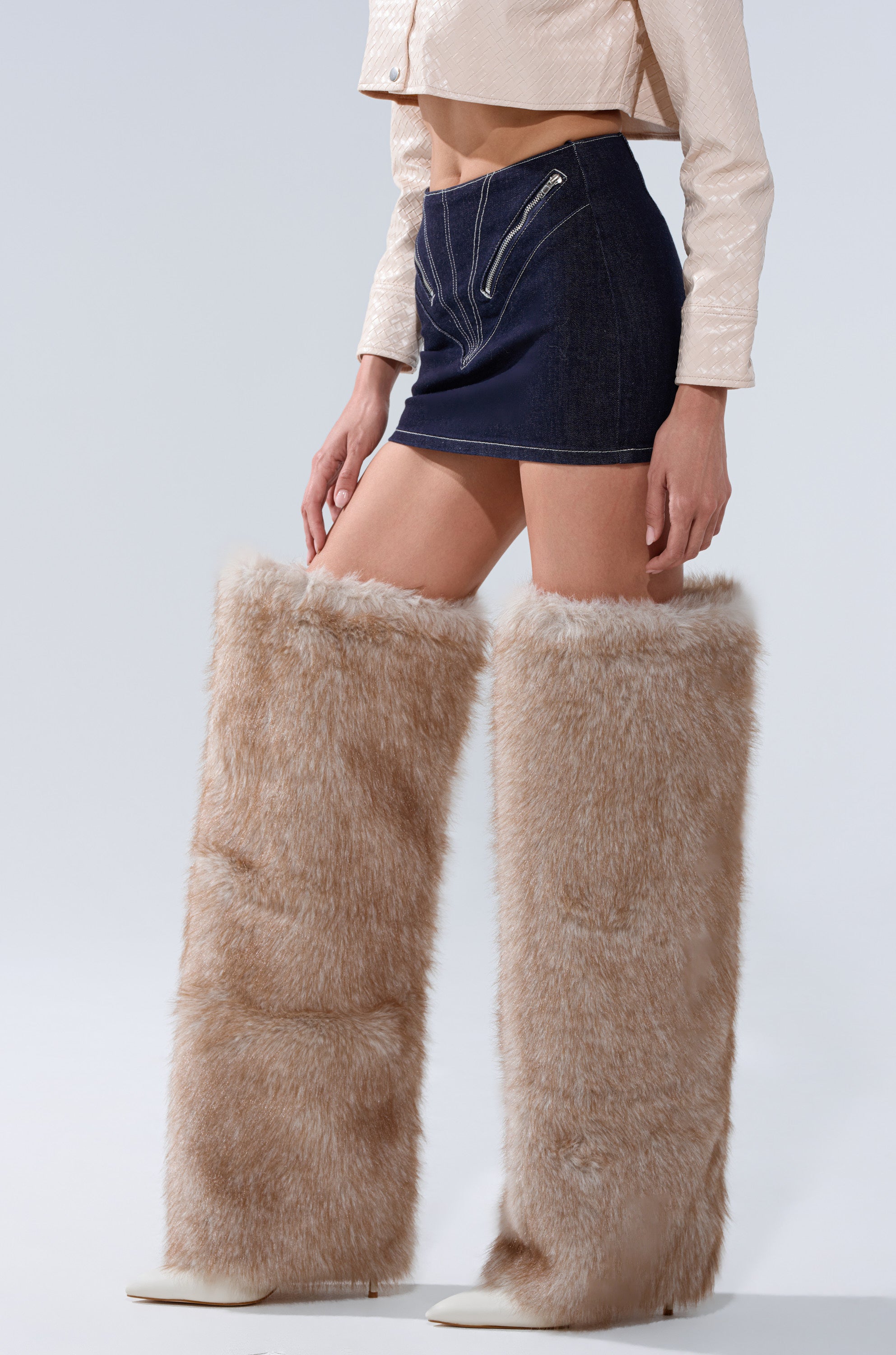 A person stands against a plain background wearing a cropped jacket, the ZIP ME UP DENIM MINI SKIRT with zippered waist pockets, and oversized tan faux fur leg warmers over white shoes. 3