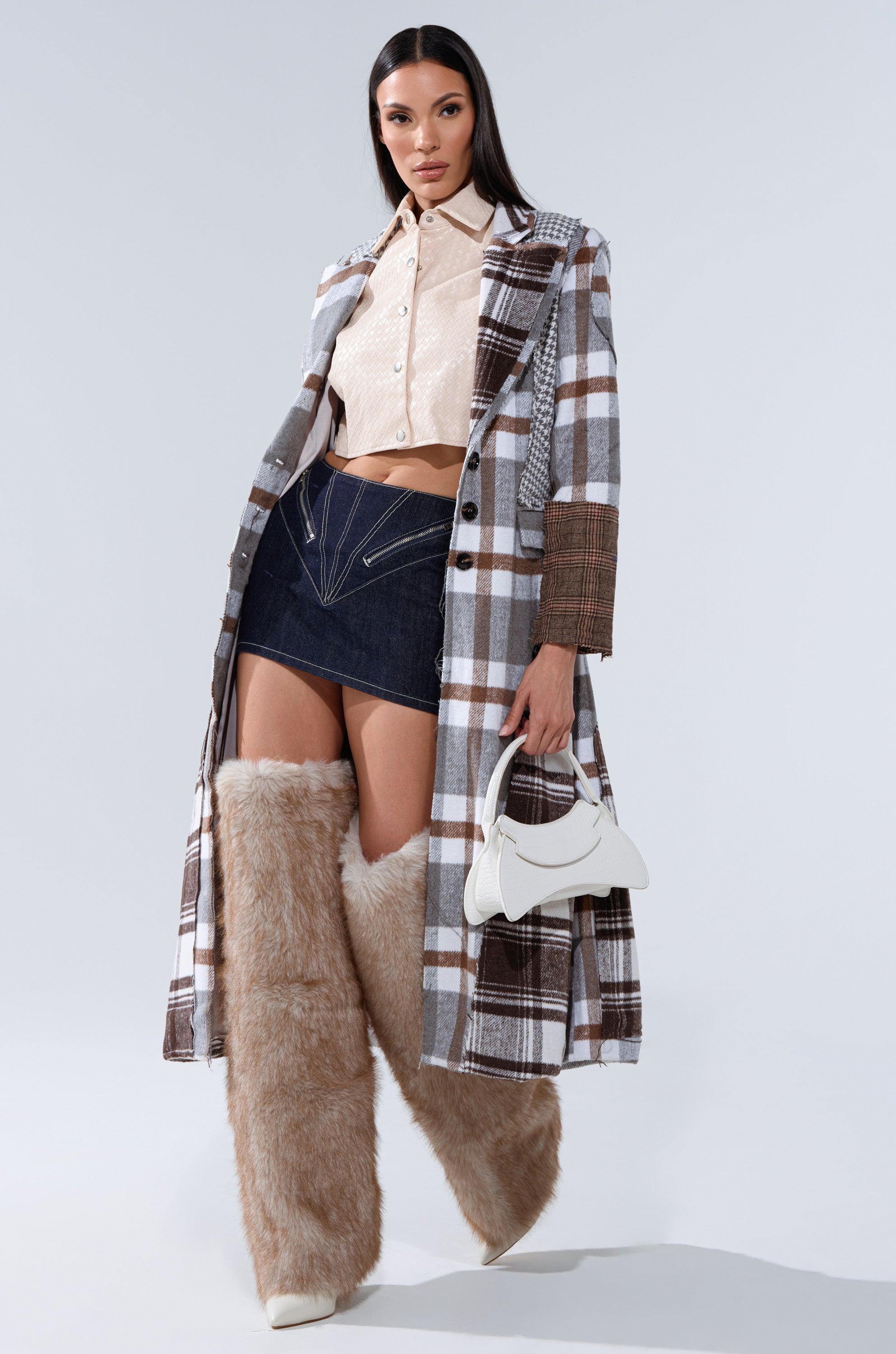 A woman poses in a long plaid coat, beige button-up shirt, ZIP ME UP DENIM MINI SKIRT, oversized furry beige leg warmers, white pointed boots, and holds a small white handbag against a plain light background. 8