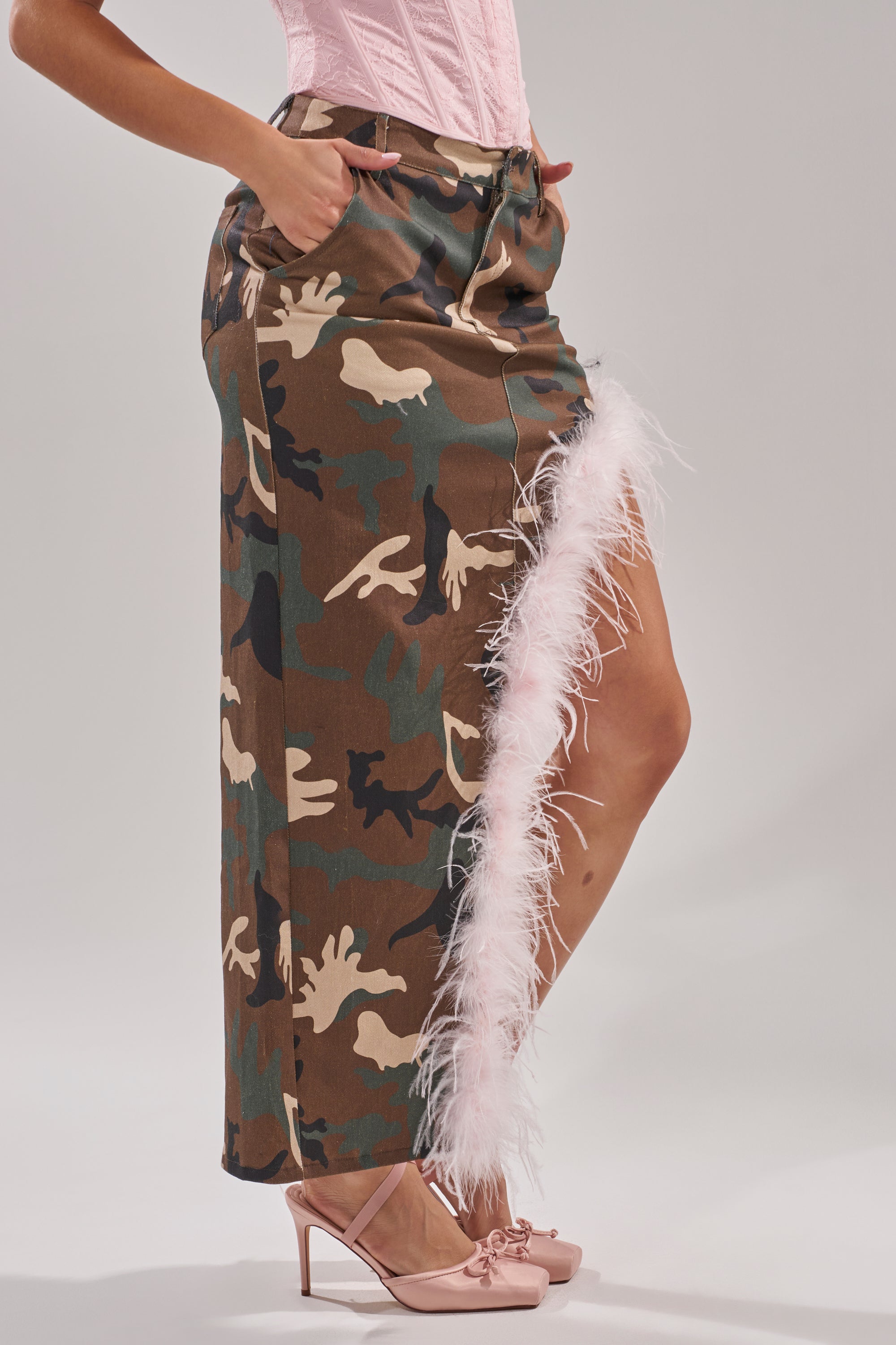 A person stands confidently with hands in pockets, wearing a pink corset, pink bow high heels, and the TRYING SOMETHING NEW CAMO SKIRT WITH FEATHERS—a maxi skirt featuring feather trim and a high slit.