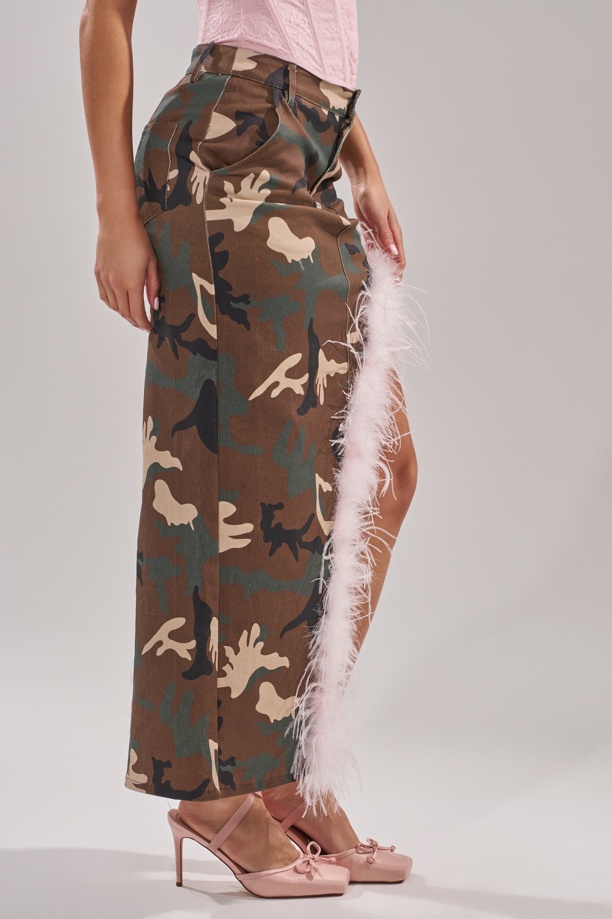 A person wearing the TRYING SOMETHING NEW CAMO SKIRT WITH FEATHERS, featuring a thigh-high slit with light pink feather trim, styled with blush pink heels and a pink top.