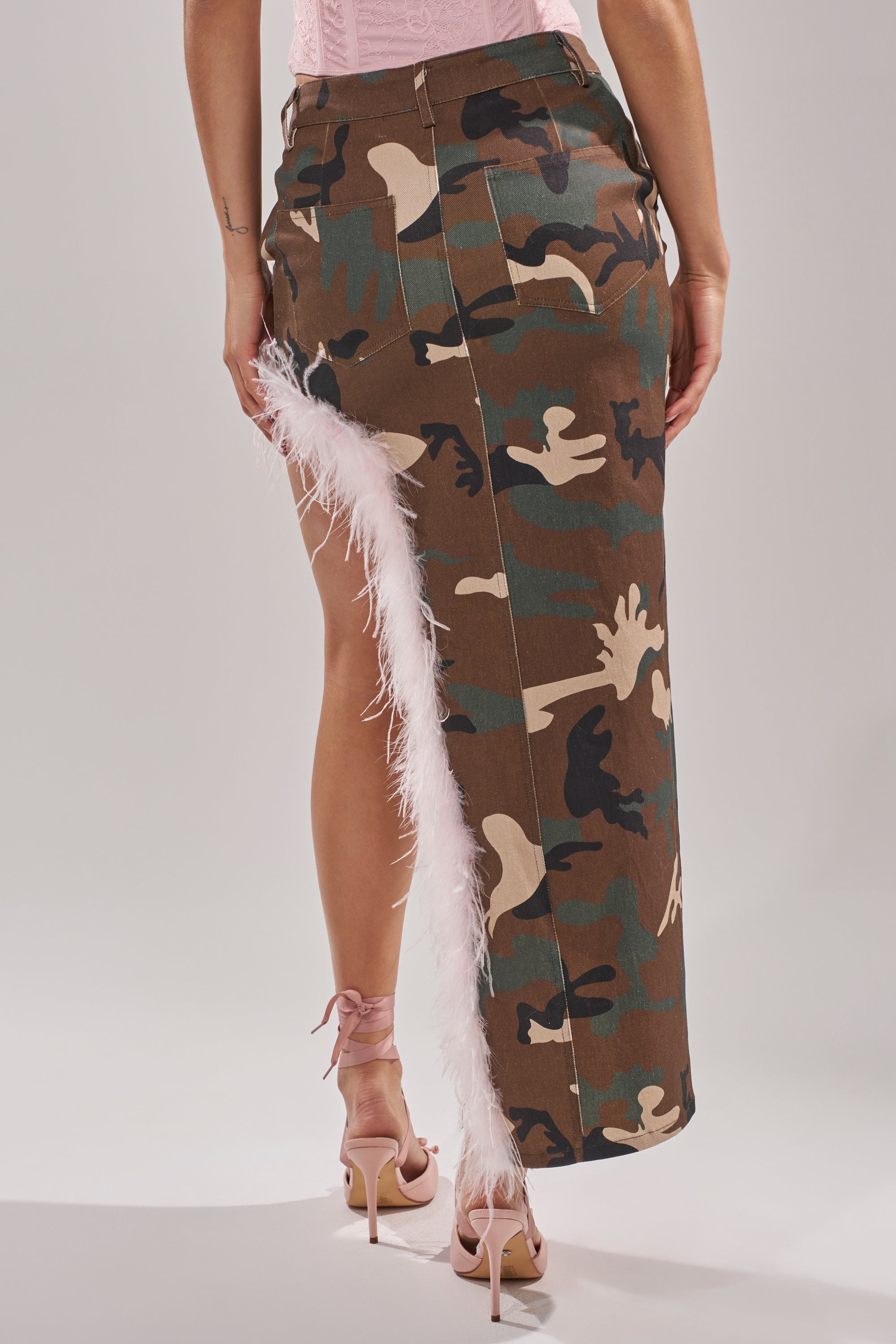 A person wears the TRYING SOMETHING NEW CAMO SKIRT WITH FEATHERS, featuring a high waist and diagonal feather-trimmed slit, paired with light pink lace-up heeled sandals. The back view highlights the skirt's unique details.
