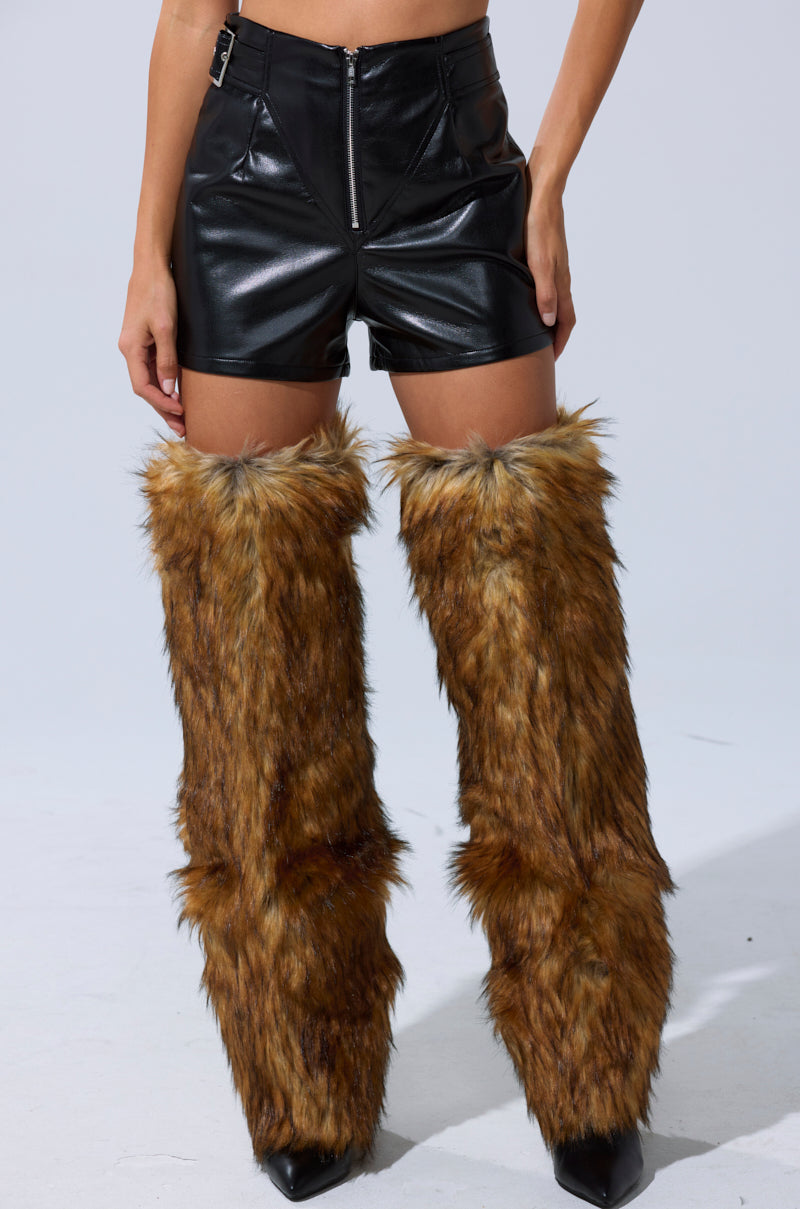 Someone wears the BUILD ME UP FAUX LEATHER SHORT—black, high-rise, with chaps-style panels—paired with brown furry leg warmers over their legs and pointed black shoes, standing against a plain light background. 2