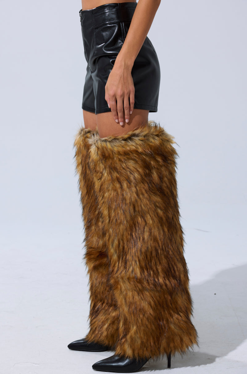 Someone wearing the BUILD ME UP FAUX LEATHER SHORT—black high-rise faux leather shorts with chaps-inspired paneling—paired with black pointed-toe heels and tall brown faux fur leg warmers, stands against a plain light background. 4